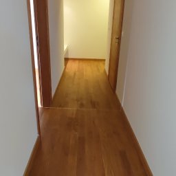 SaVo - Korytarz z drewnianą podłogą w odcieniach brązu, świeżo po remoncie, z widocznymi dwiema parami drzwi i jasnym pomieszczeniem w głębi.