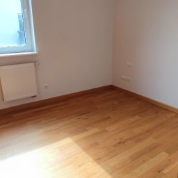 SaVo - Pusty pokój z drewnianą podłogą, świeżo pomalowanymi białymi ścianami, grzejnikiem pod oknem i widocznymi gniazdkami elektrycznymi, sfotografowany zza częściowo otwartych drewnianych drzwi.