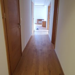 SaVo - Długi korytarz z drewnianą podłogą i drzwiami po obu stronach, prowadzący do jasnego pomieszczenia z biurkiem i szafką.