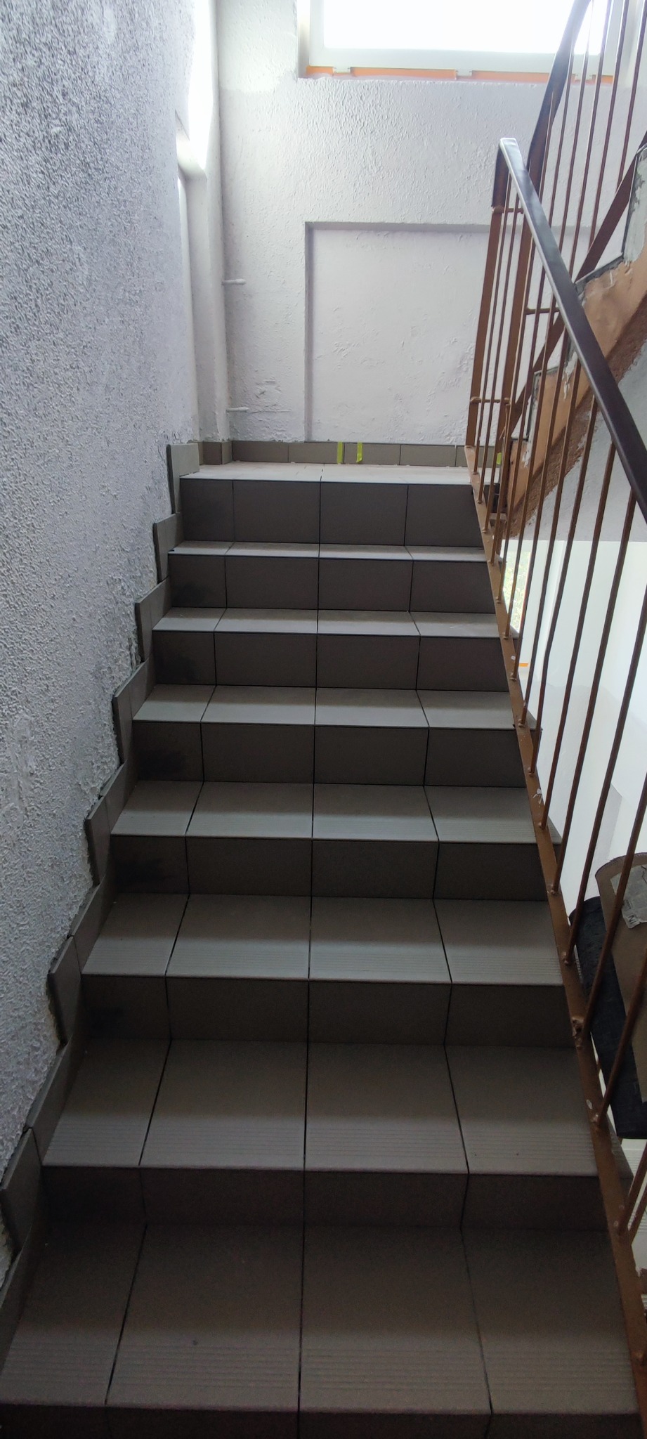 Klatka schodowa wyłożona szarymi płytkami, widok z dołu na górę, z metalową poręczą po prawej stronie i ścianą o szorstkiej fakturze po lewej.