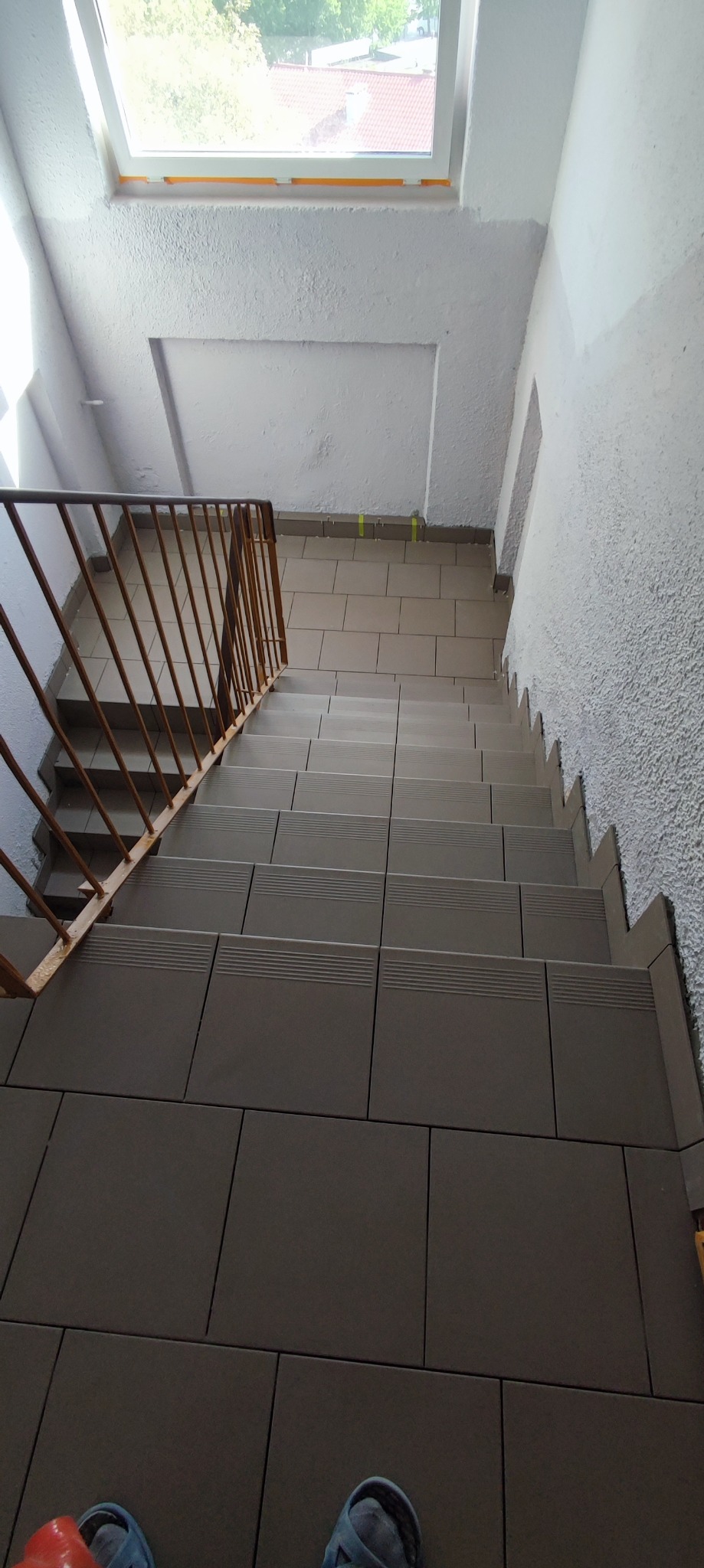 Klatka schodowa w trakcie remontu, widok z góry na wyłożone płytkami schody i pomalowane ściany z oknem na górze, zabezpieczone taśmą malarską.