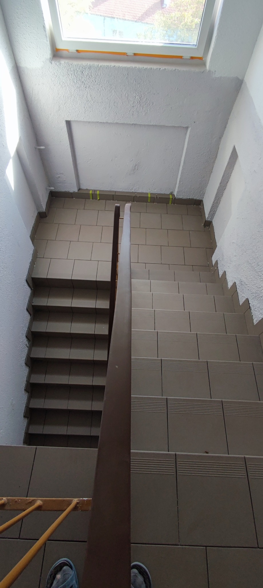 Klatka schodowa w trakcie wykańczania, widok z góry na brązowe płytki na schodach i żółtą taśmę maskującą przy suficie, drewniana poręcz i fragment stopy w klapku.