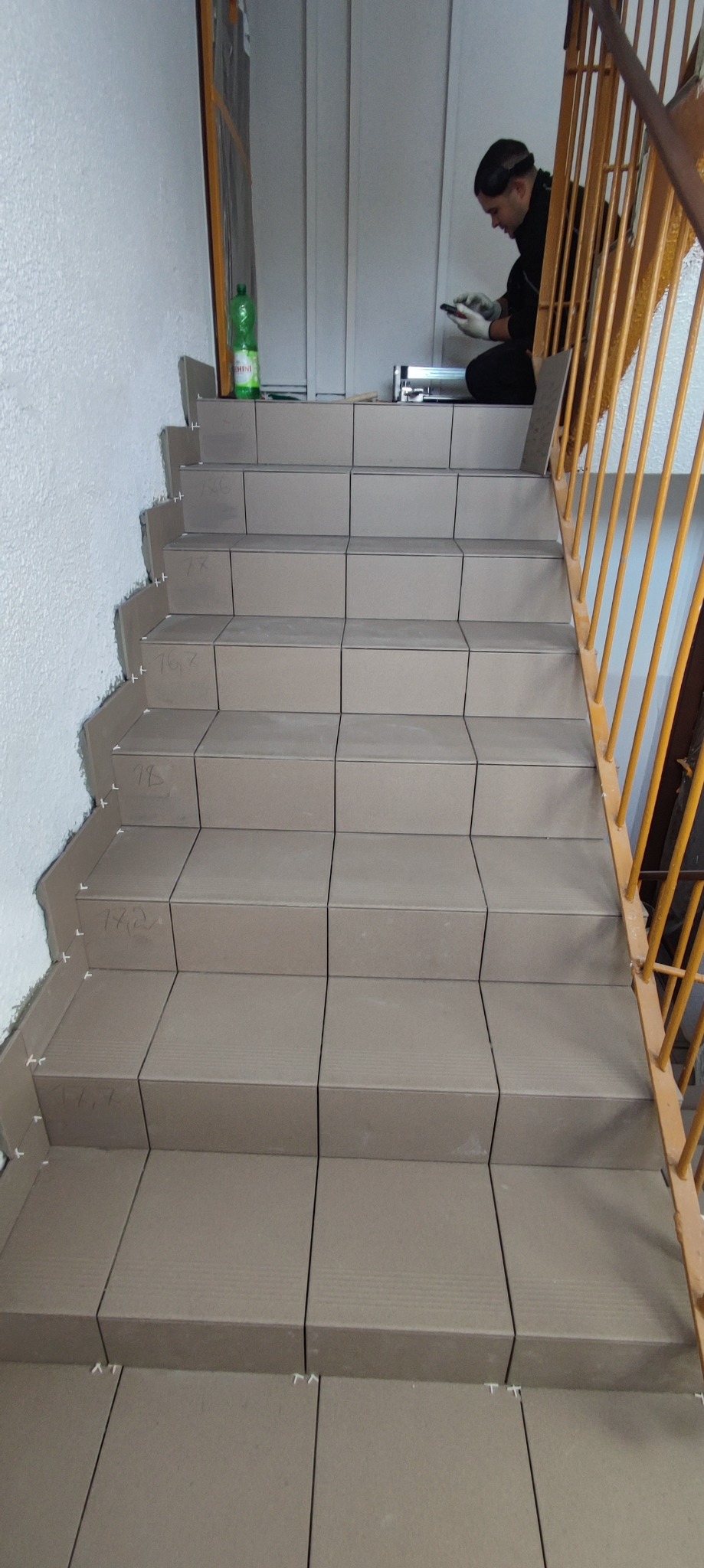 Układanie szarych płytek na schodach w klatce schodowej, widoczne plastikowe krzyżyki dystansowe, butelka wody i pracownik w tle.