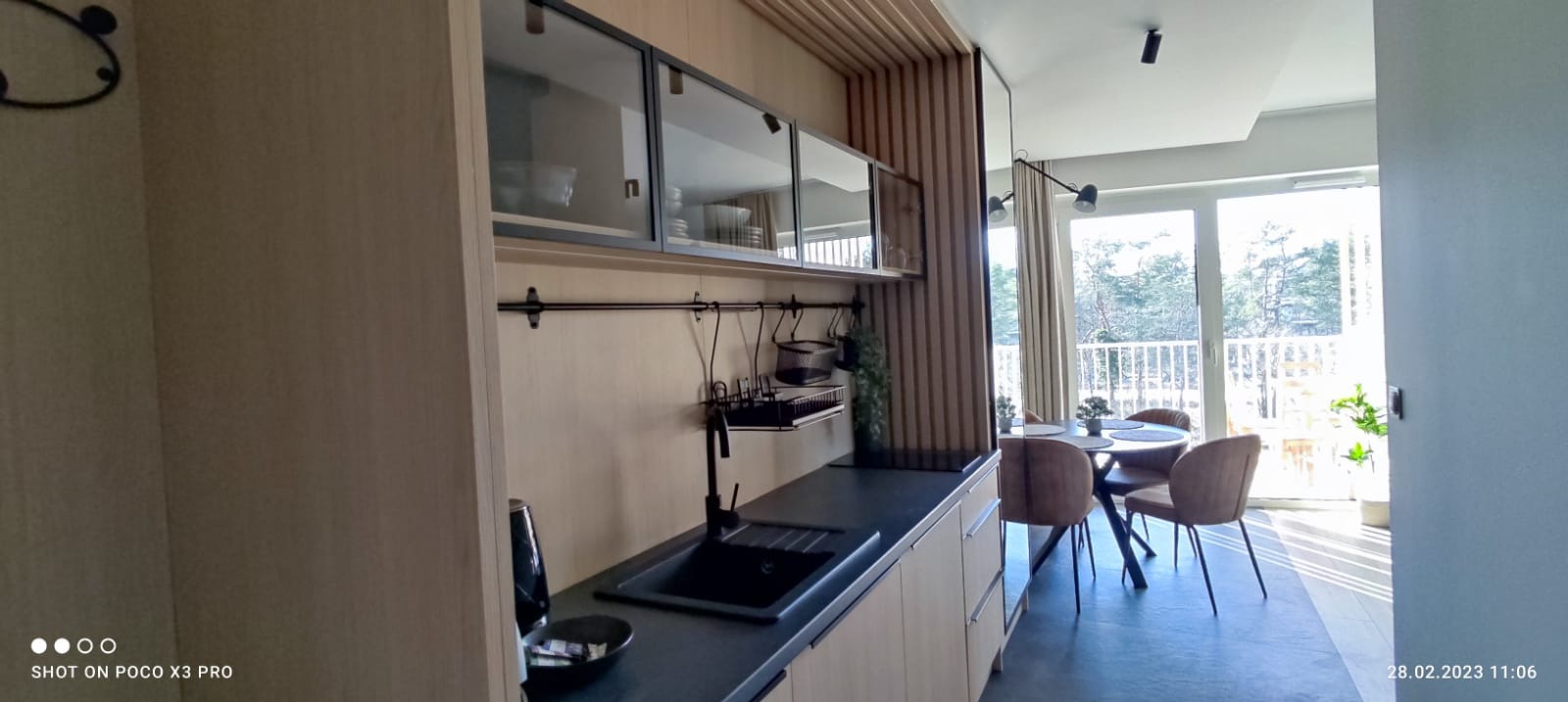 Jasna kuchnia z drewnianymi akcentami, czarnym zlewem i przeszklonymi szafkami wiszącymi; widok na stół z krzesłami i balkon z zielenią w tle.