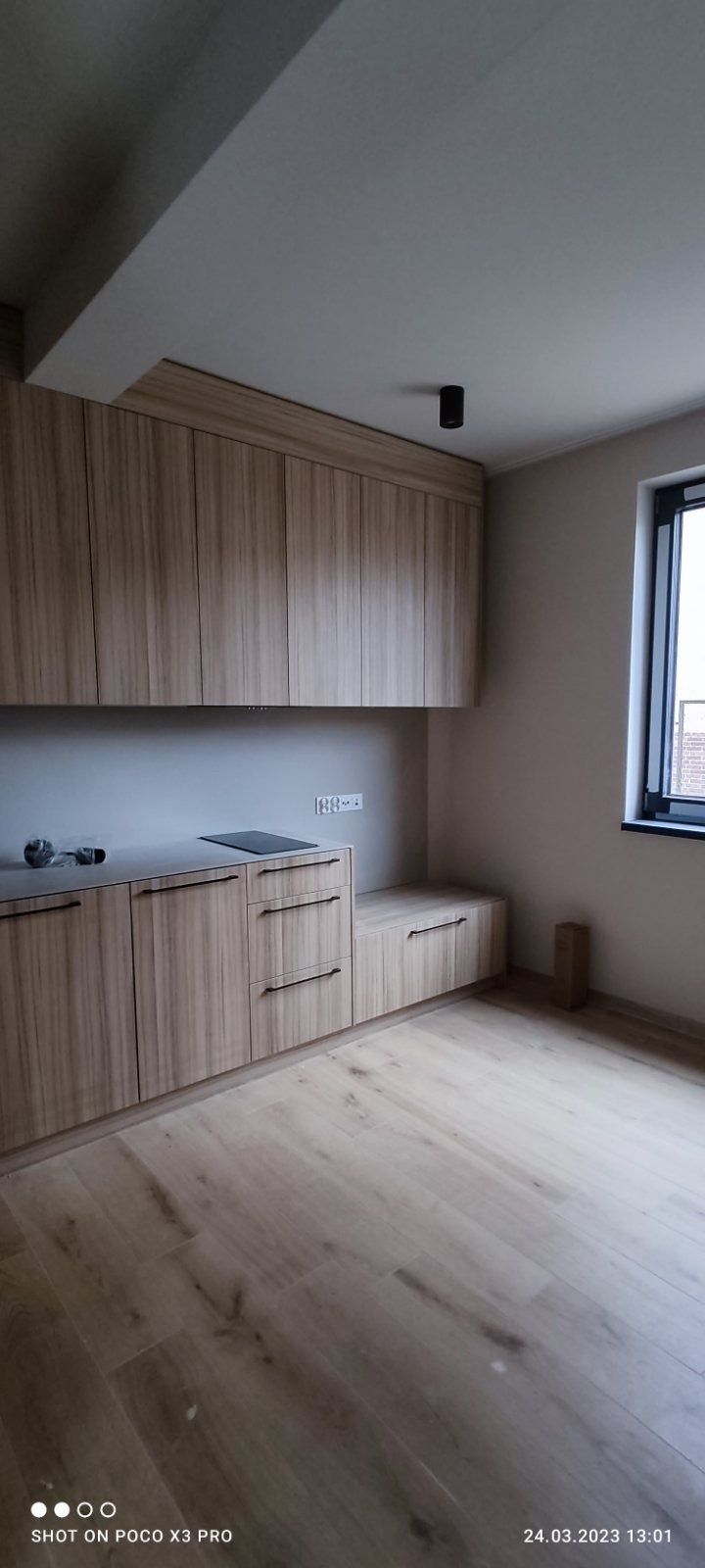 Nowoczesna kuchnia z drewnianymi szafkami, czarnymi uchwytami i płytą indukcyjną, widok na okno z czarną ramą, minimalistyczne oświetlenie sufitowe.