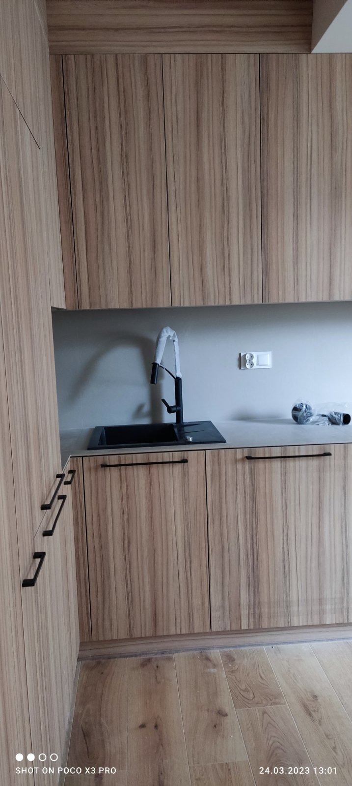 Nowoczesna zabudowa kuchenna z drewnopodobnymi frontami, czarnym zlewem i baterią, widoczne akcesoria montażowe w folii, jeszcze przed pełnym uruchomieniem.