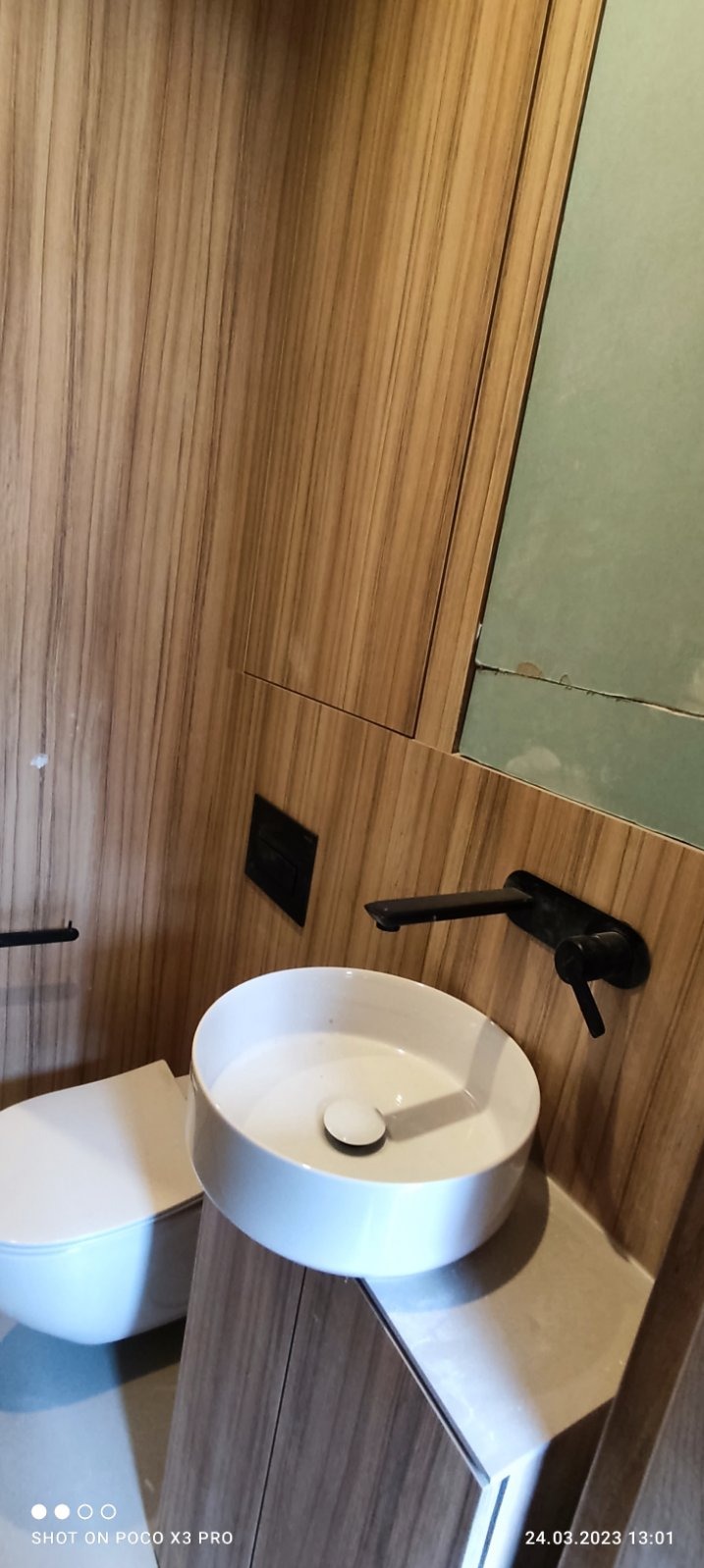 Nowoczesna łazienka w trakcie wykańczania: biała, okrągła umywalka nablatowa z czarną armaturą, drewnopodobne panele ścienne i fragment surowej ściany przygotowanej do wykończenia.