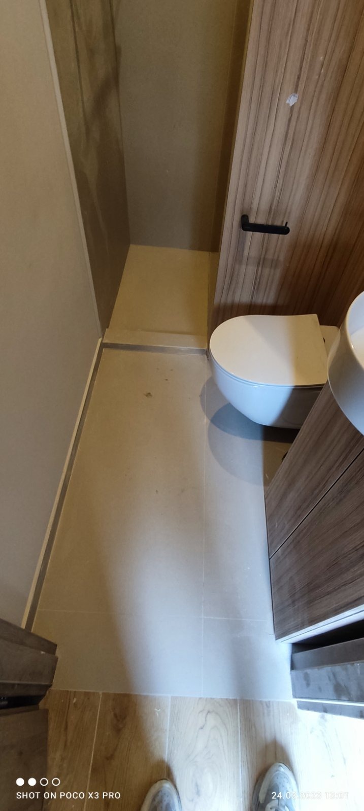 Nowoczesna łazienka po remoncie z beżowymi płytkami podłogowymi i ściennymi, podwieszana toaleta oraz umywalka z szafką z drewnopodobnym wykończeniem, widoczne buty osoby robiącej zdjęcie.