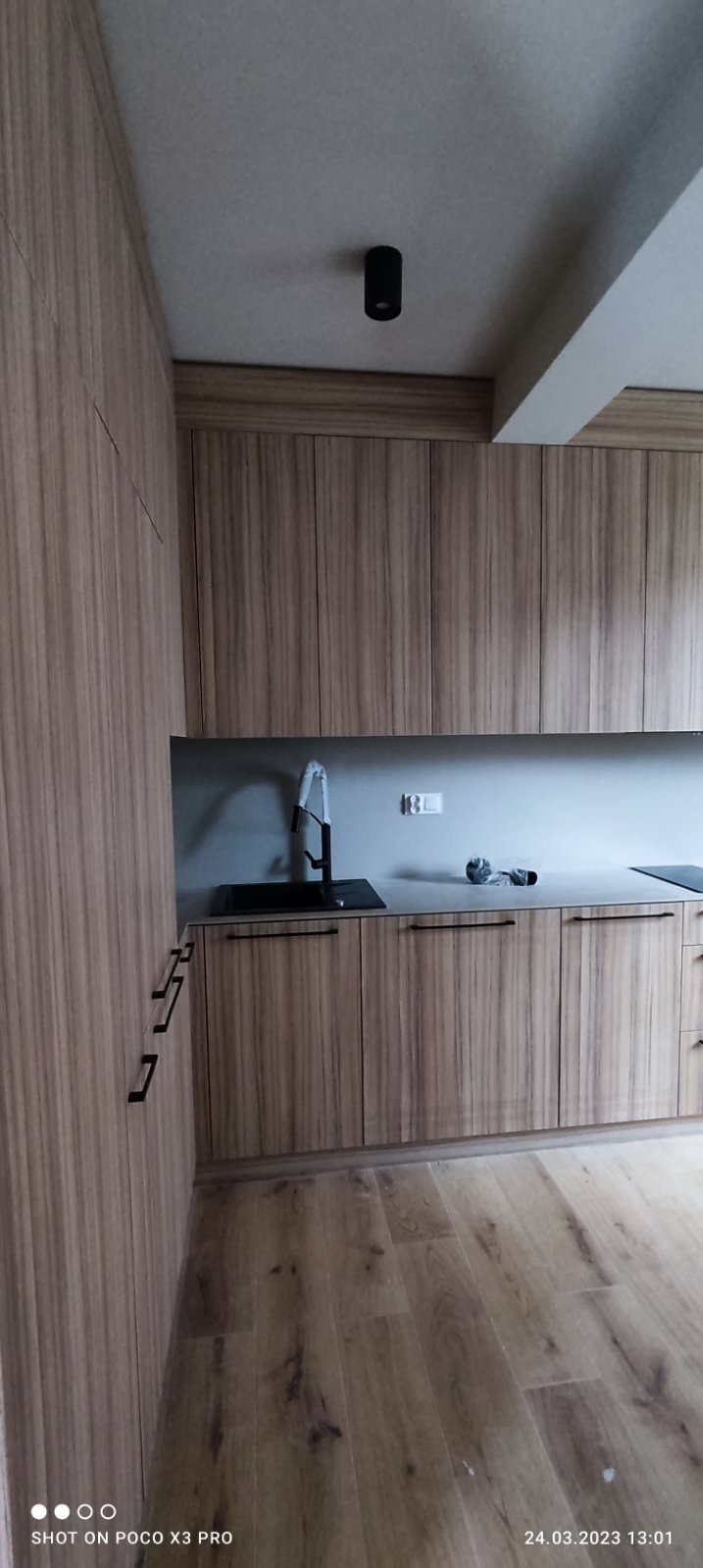 Nowoczesna kuchnia z drewnopodobnymi frontami szafek, czarną armaturą i jasną podłogą laminowaną. Widoczny minimalistyczny czarny punkt świetlny na suficie.