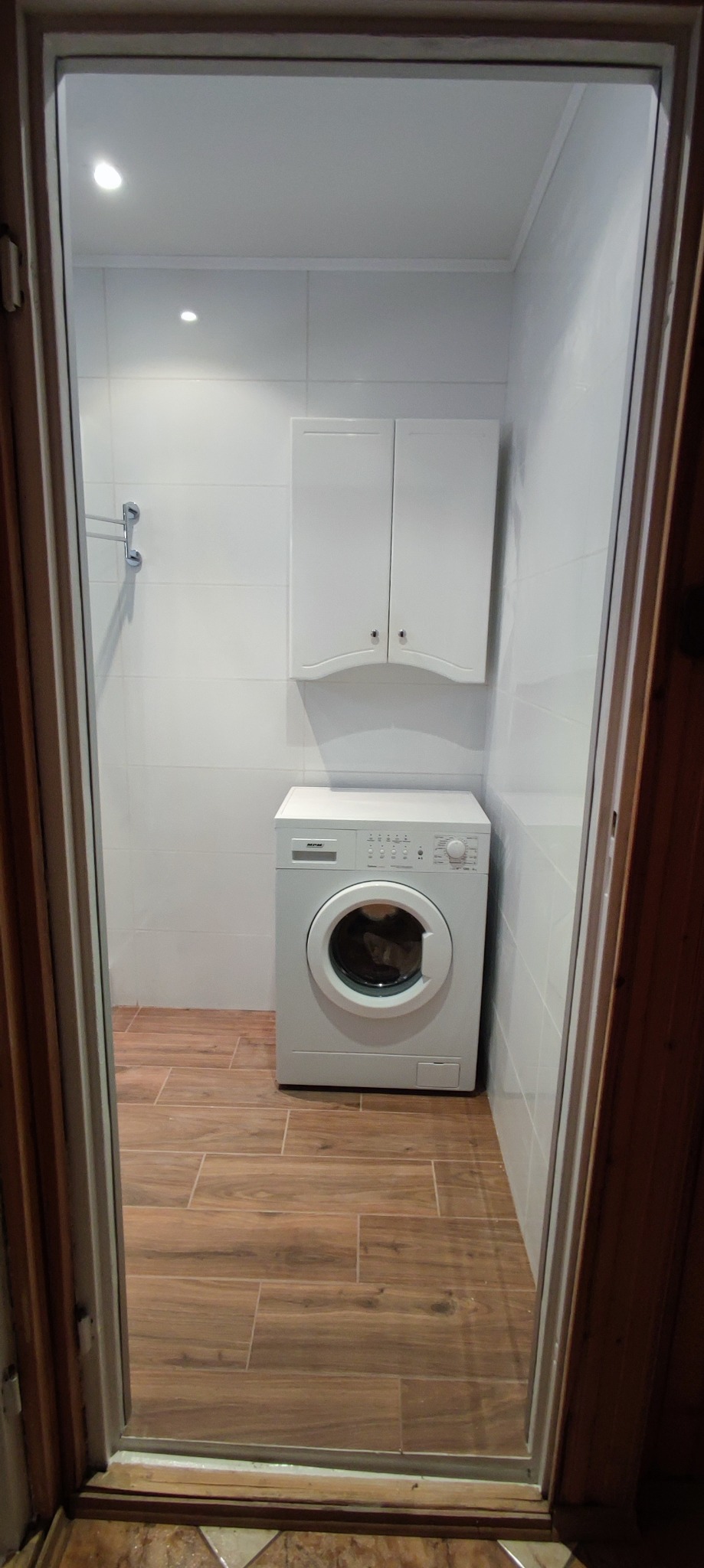 Umywalka z szafką i pralką w wyremontowanej łazience z brązowymi płytkami podłogowymi imitującymi drewno, białe płytki ścienne, widok z korytarza.