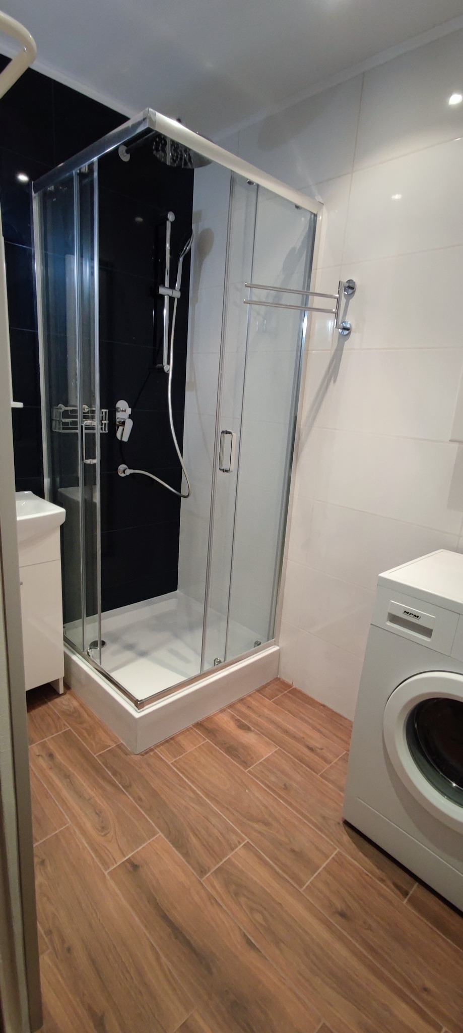 Nowoczesna łazienka w Szczecinie z kabiną prysznicową typu walk-in na tle czarnych płytek, umywalką z szafką i pralką, wykończona drewnopodobną podłogą.