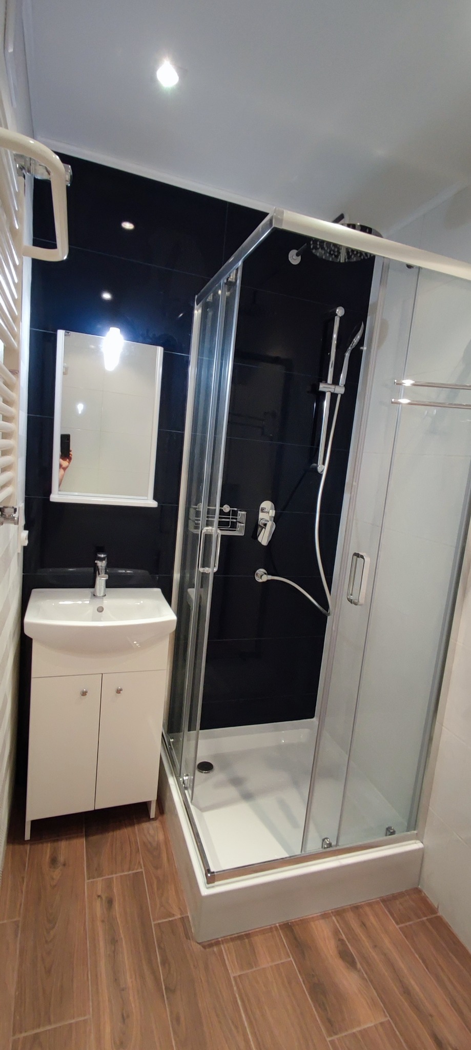 Nowoczesna łazienka z czarnymi płytkami na ścianie, kabiną prysznicową z przezroczystego szkła i białą szafką pod umywalką, uzupełniona drewnopodobną podłogą i grzejnikiem drabinkowym.