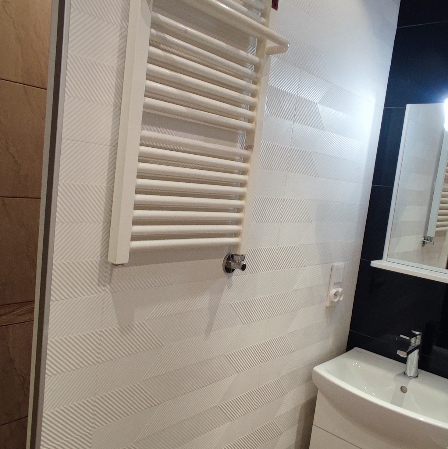 Biała łazienka z grzejnikiem drabinkowym na ścianie z wzorzystych płytek, umywalka z baterią i fragment lustra z czarną ramą.