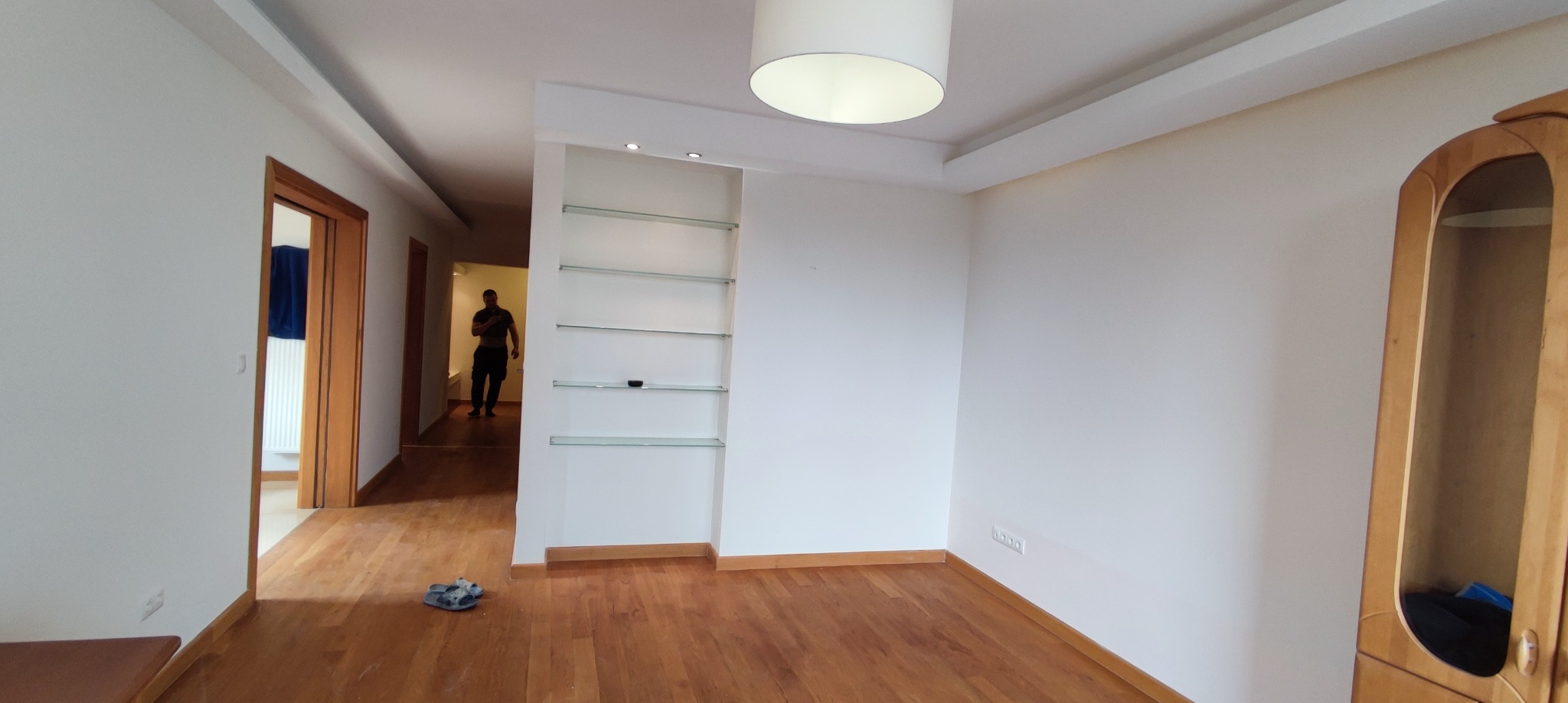 Pusta, jasna przestrzeń pokoju z drewnianą podłogą, białymi ścianami, wbudowanymi szklanymi półkami i widoczną osobą w przejściu do kolejnego pomieszczenia.