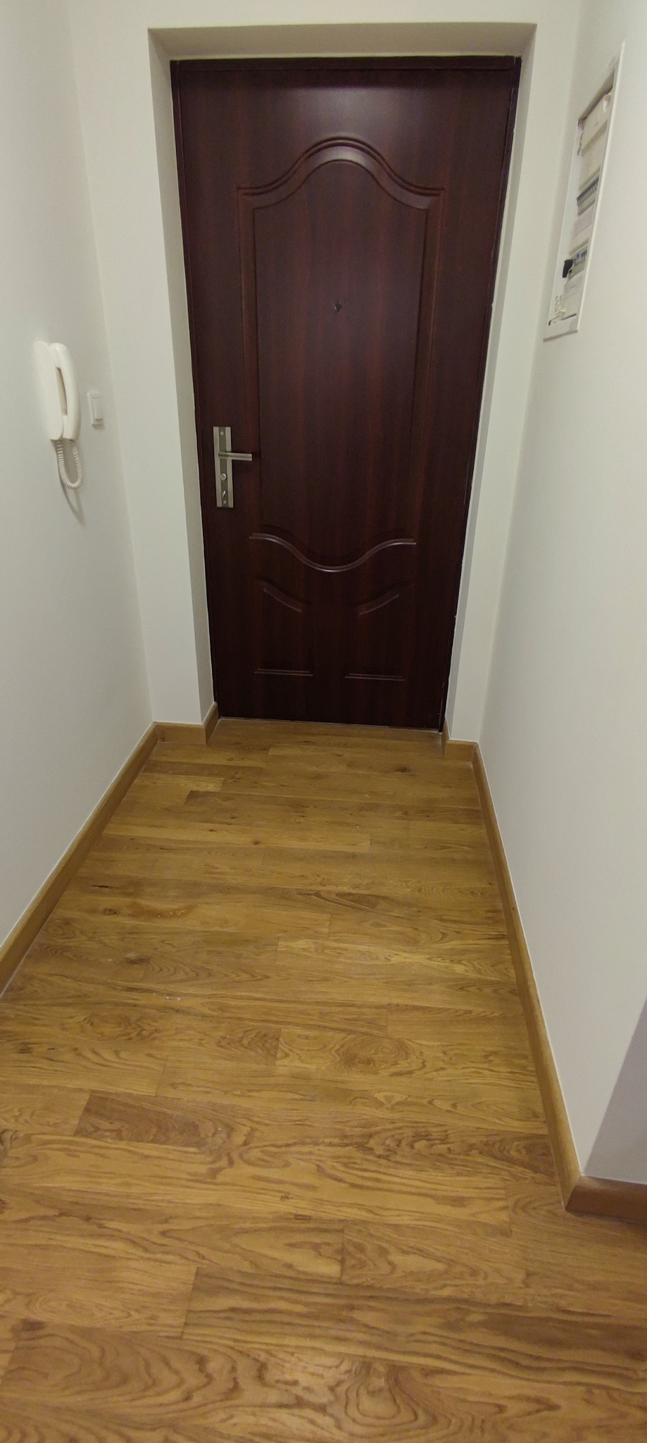 Widok na świeżo położoną podłogę z paneli imitujących drewno, prowadzącą do ciemnobrązowych drzwi wejściowych w białym korytarzu. Na ścianie widoczny biały domofon i skrzynka elektryczna.