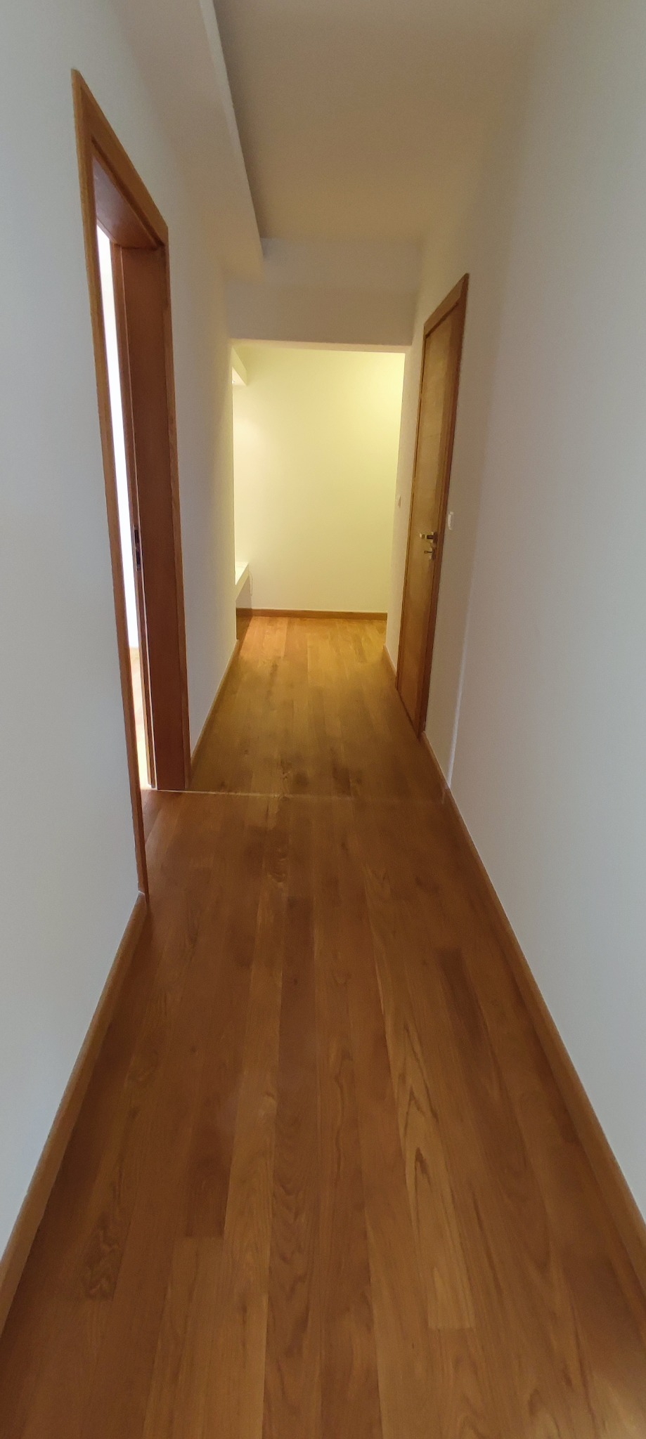 Korytarz z drewnianą podłogą w odcieniach brązu, świeżo po remoncie, z widocznymi dwiema parami drzwi i jasnym pomieszczeniem w głębi.