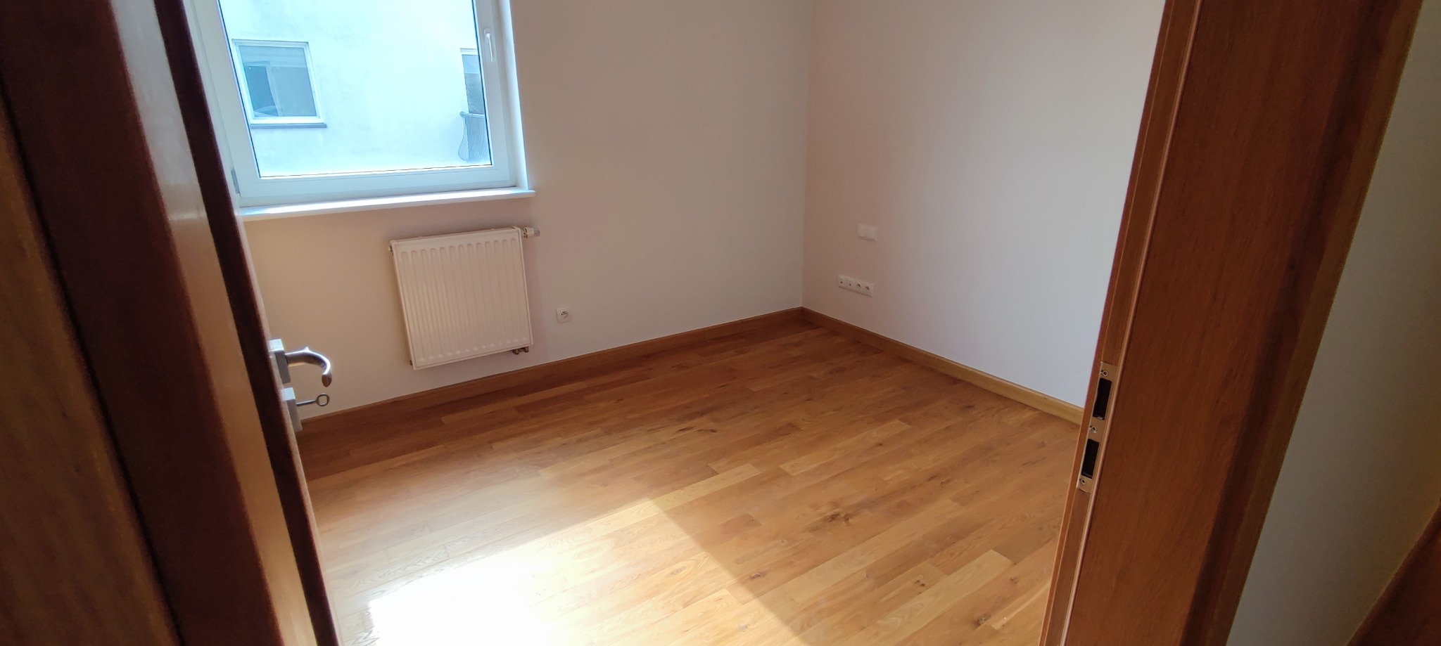 Pusty pokój z drewnianą podłogą, świeżo pomalowanymi białymi ścianami, grzejnikiem pod oknem i widocznymi gniazdkami elektrycznymi, sfotografowany zza częściowo otwartych drewnianych drzwi.