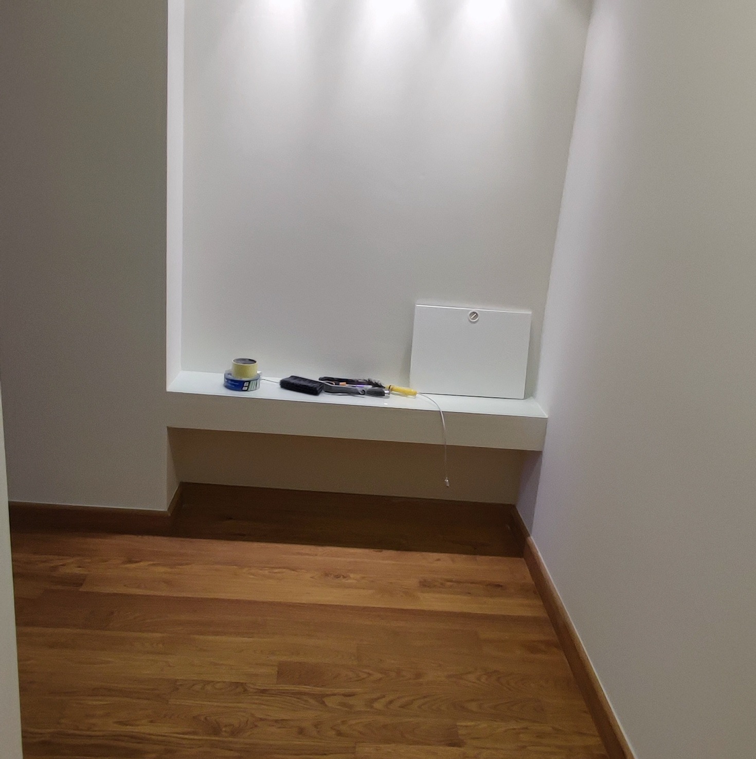 Wnęka w świeżo pomalowanej ścianie z półką, na której leżą narzędzia, taśma malarska i skrzynka rewizyjna. Parkiet na podłodze.
