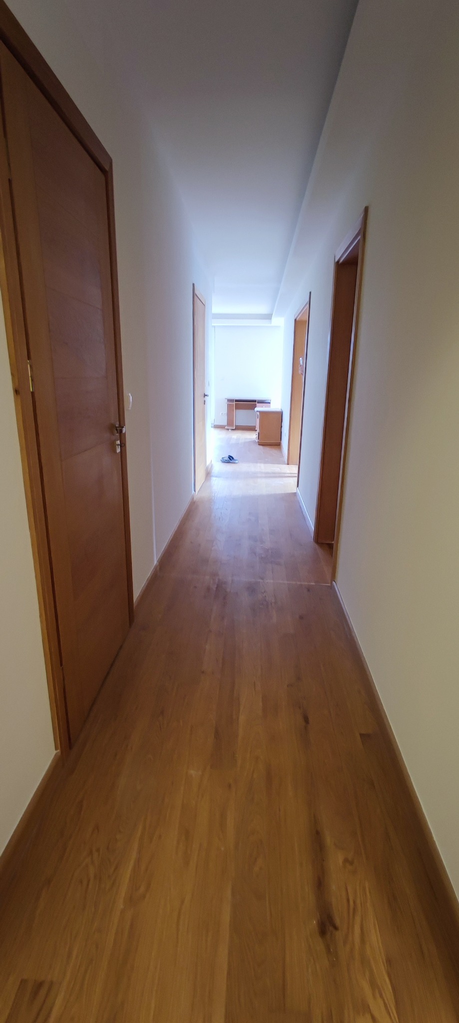 Długi korytarz z drewnianą podłogą i drzwiami po obu stronach, prowadzący do jasnego pomieszczenia z biurkiem i szafką.