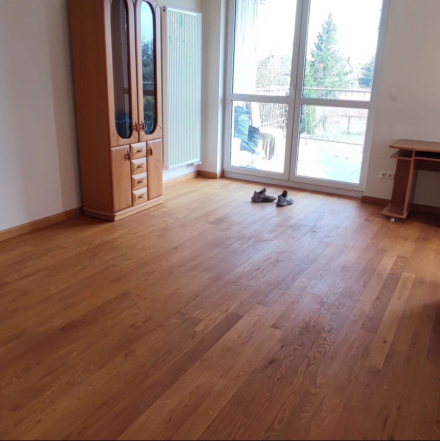 Pusty pokój po remoncie z nową, drewnianą podłogą, widoczne buty przy oknie, grzejnik i drewniany kredens.