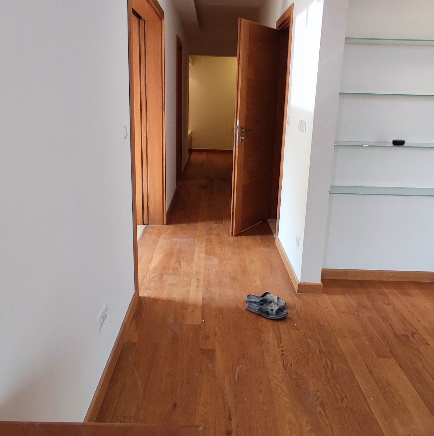 Wnętrze mieszkania po remoncie: parkiet z drewna egzotycznego, białe ściany, drewniane drzwi wewnętrzne, widoczne półki szklane i para klapek leżąca na podłodze.