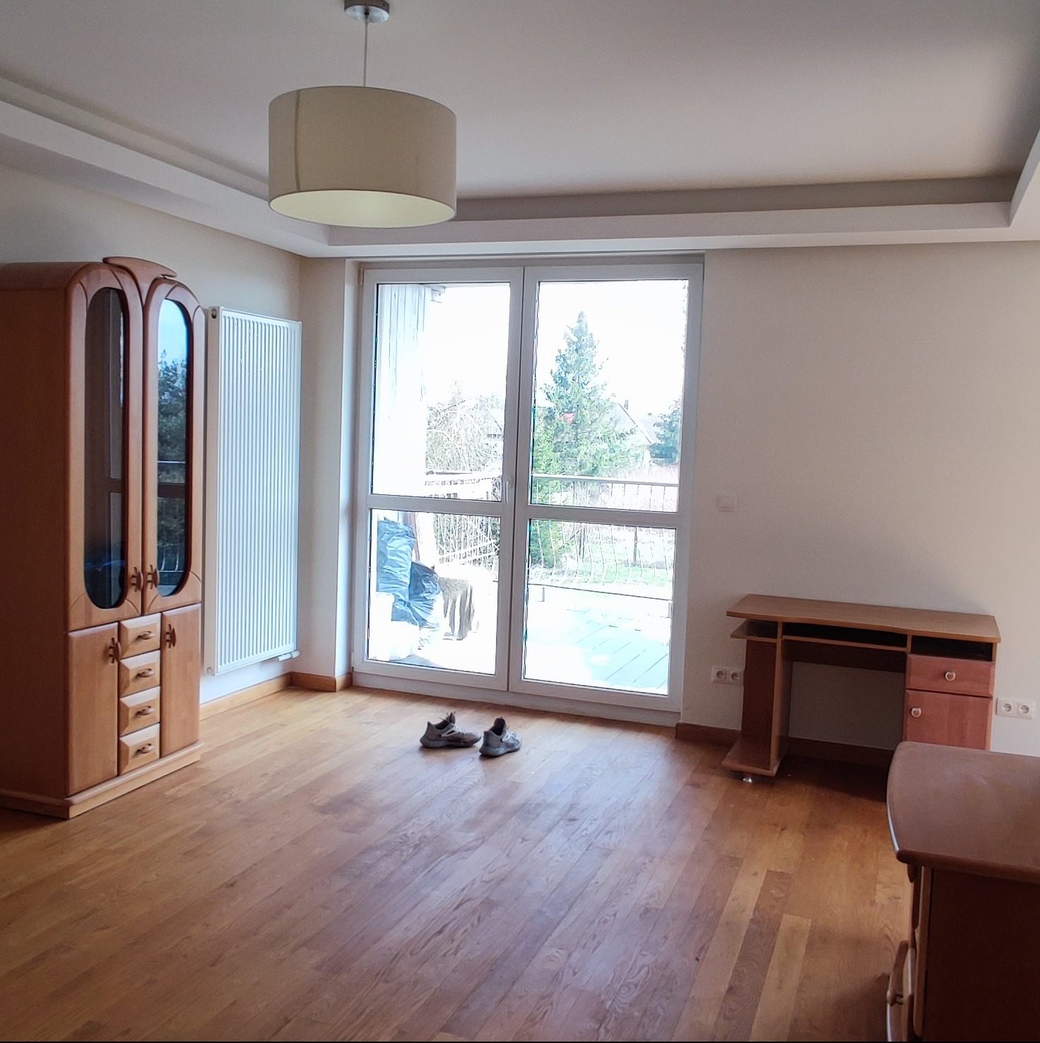 Jasne, przestronne wnętrze pokoju z drewnianą podłogą, meblami w stylu retro i dużym oknem balkonowym z widokiem na zieleń. Na podłodze leżą buty, a na suficie wisi okrągła lampa.