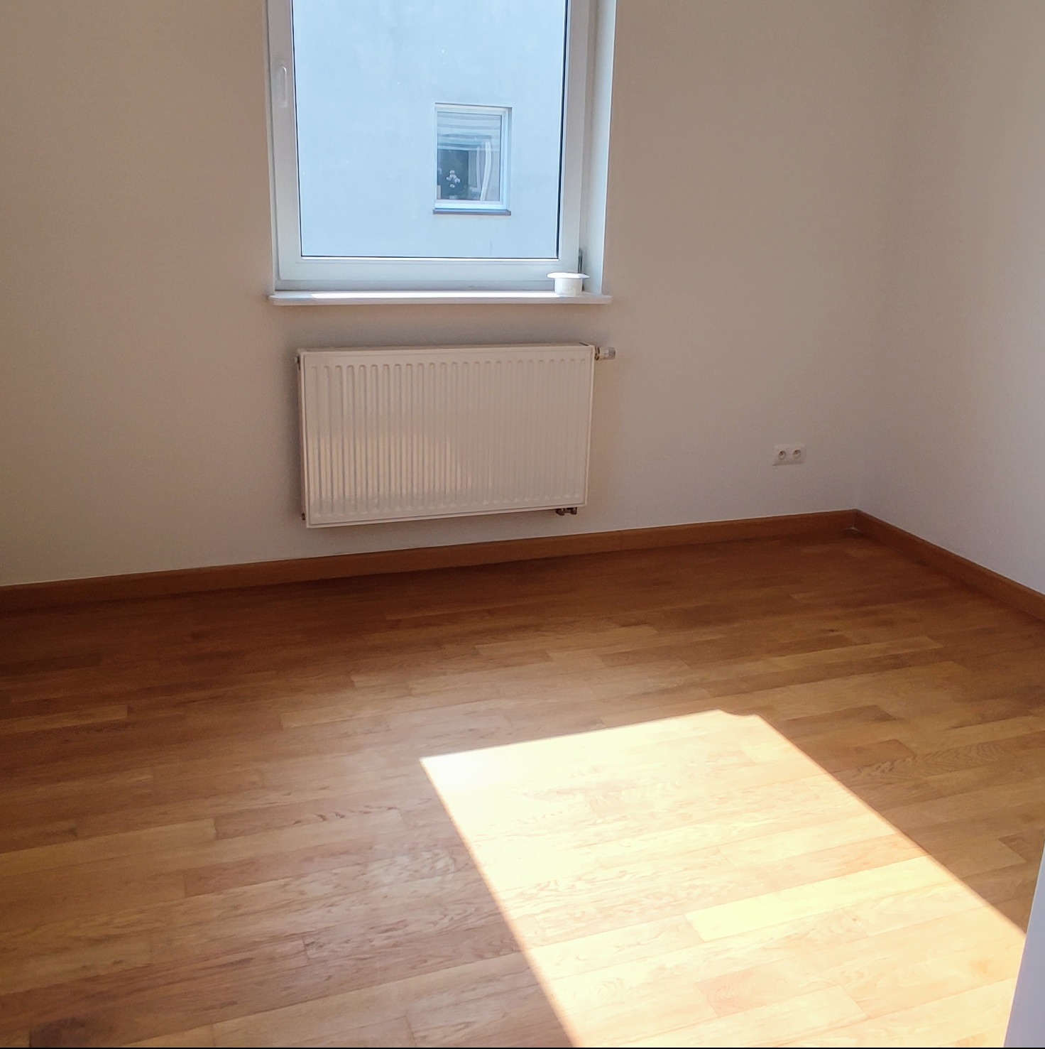 Pusty pokój z drewnianą podłogą, białymi ścianami, grzejnikiem pod oknem i silnym światłem słonecznym padającym na podłogę.