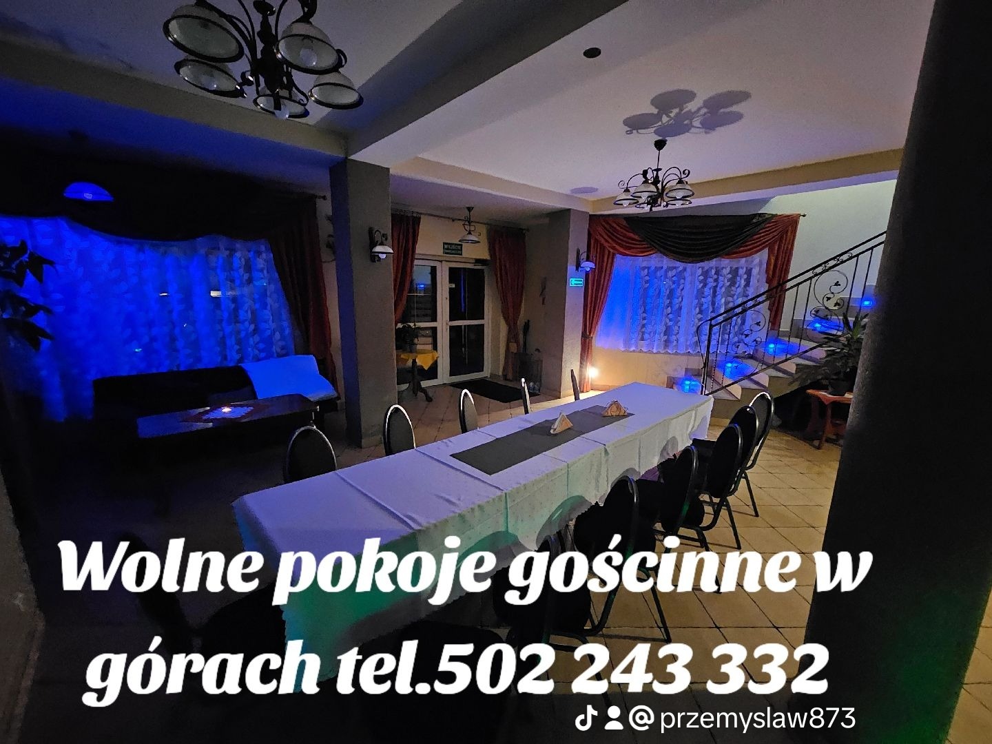 Wnętrze pokoju gościnnego z długim stołem nakrytym białym obrusem i czarnymi krzesłami, oświetlone niebieskim światłem, widoczne schody z podświetleniem.