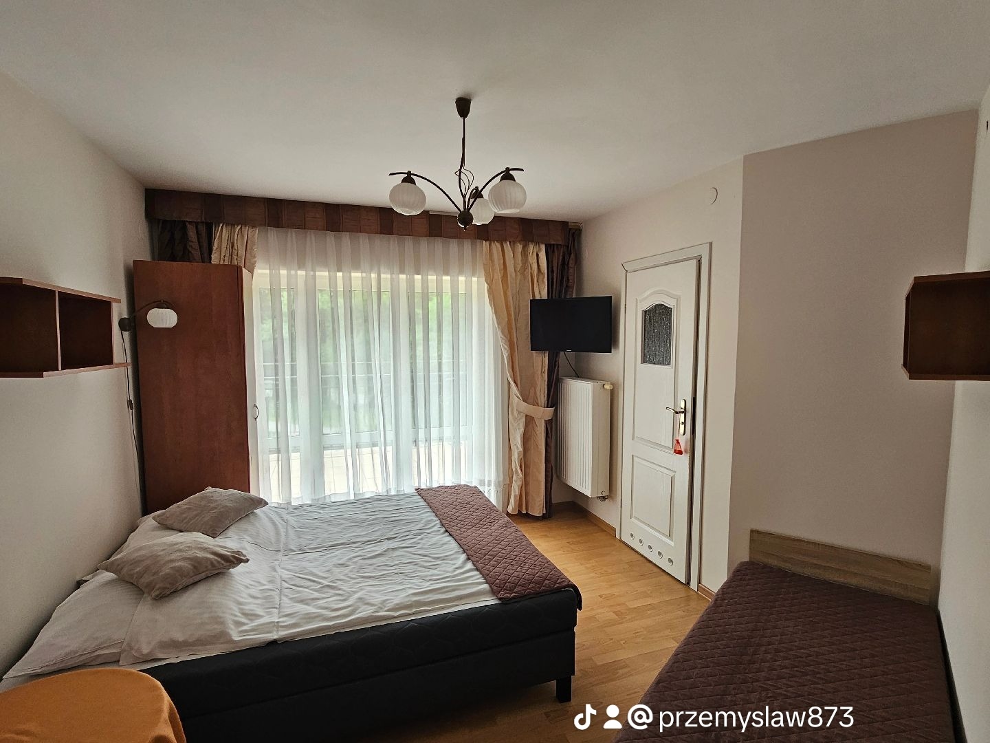 Pokój hotelowy z dwoma łóżkami, telewizorem na ścianie i oknem z zasłonami, widok z niskiego kąta.
