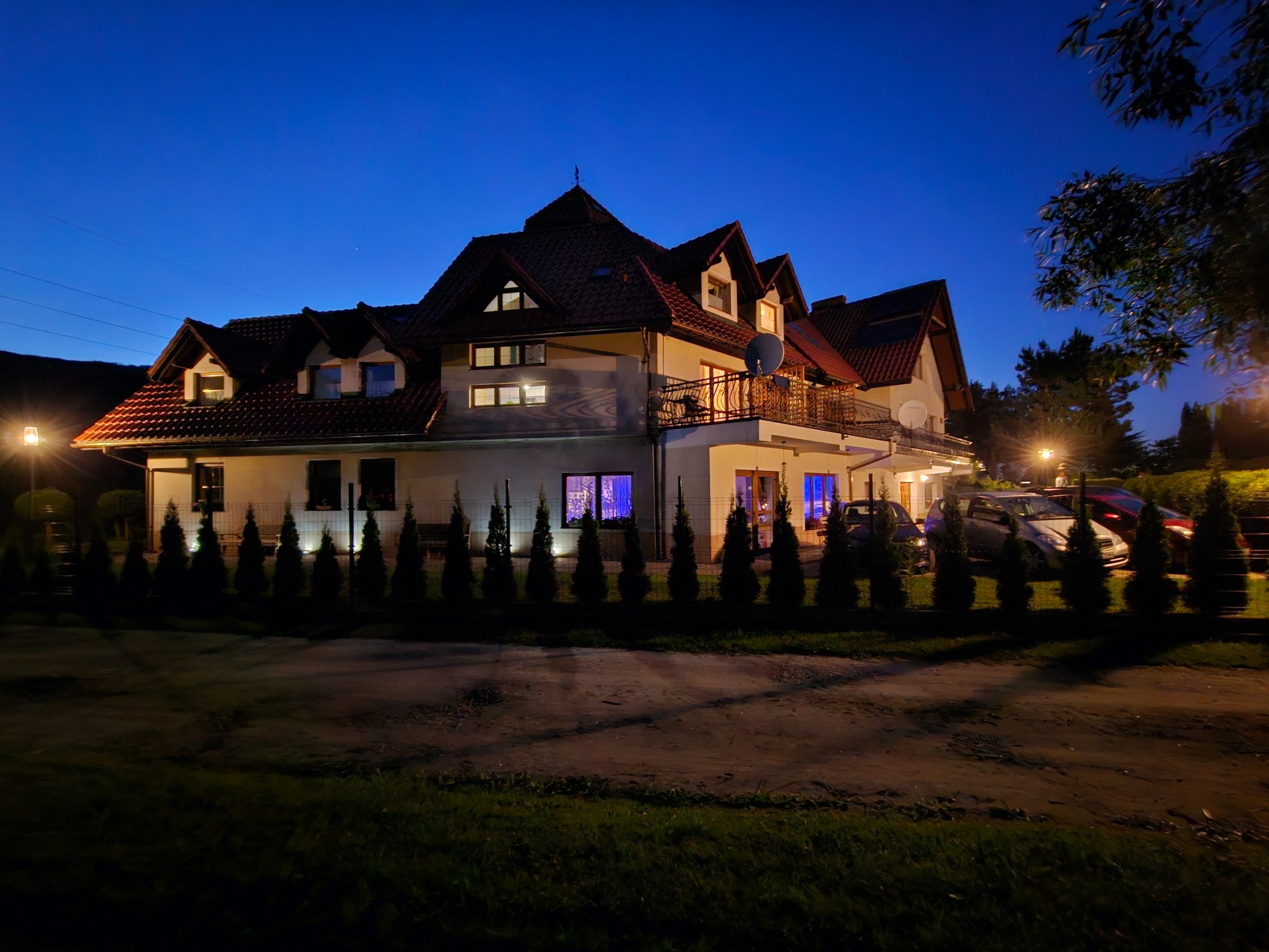 Dwupiętrowy dom z balkonem i czerwonym dachem oświetlony nocą, z rzędem małych drzew iglastych przed budynkiem i zaparkowanymi samochodami w tle.