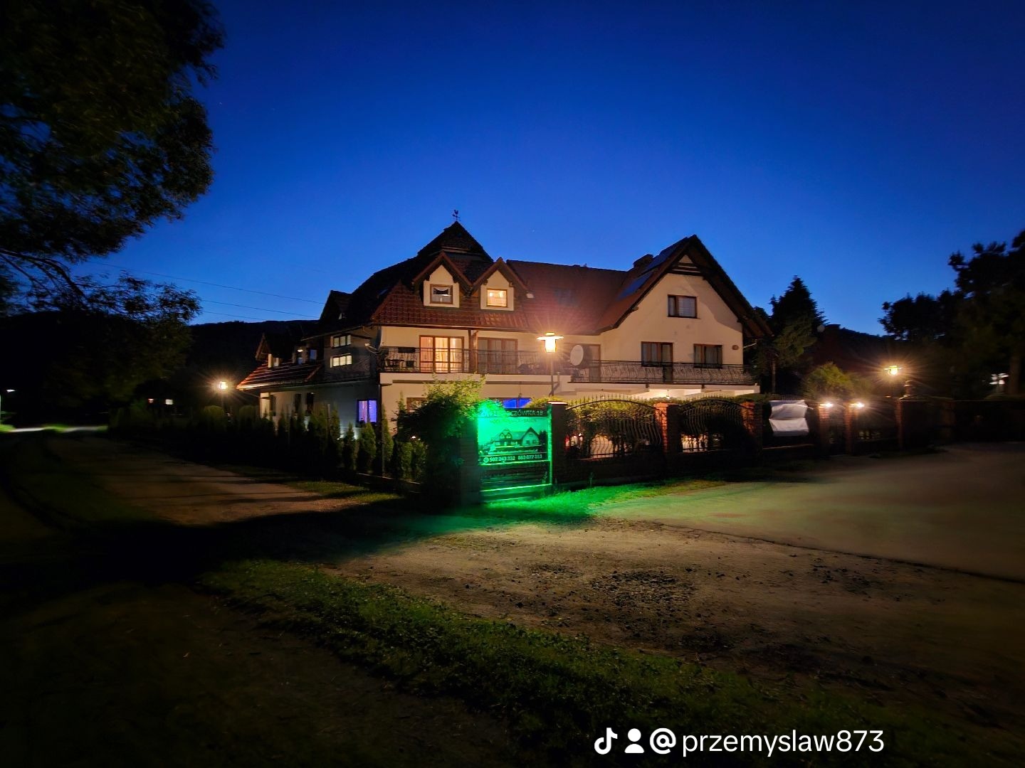 Dwupiętrowy pensjonat z balkonami oświetlony lampami ulicznymi i zielonym neonem w nocy.