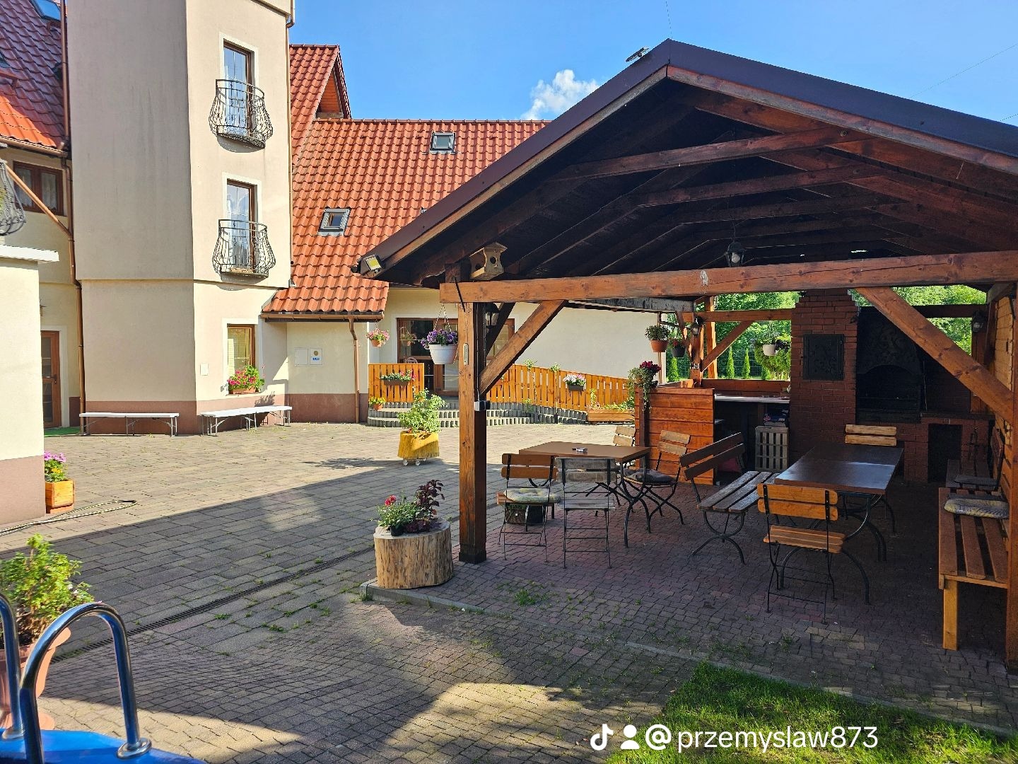 Dziedziniec z budynkiem w tle, drewnianą altaną z grillem i stołami, oraz dekoracyjnymi donicami z kwiatami.