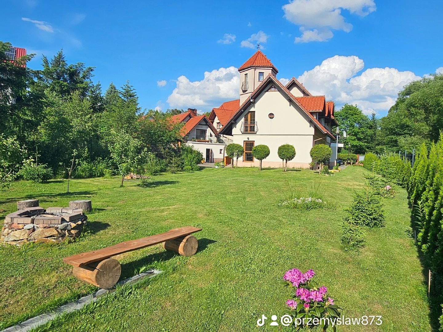 Ogród z domem jednorodzinnym w tle, drewniana ławka z okrągłymi nogami na pierwszym planie, kwitnące rododendrony w prawym dolnym rogu.