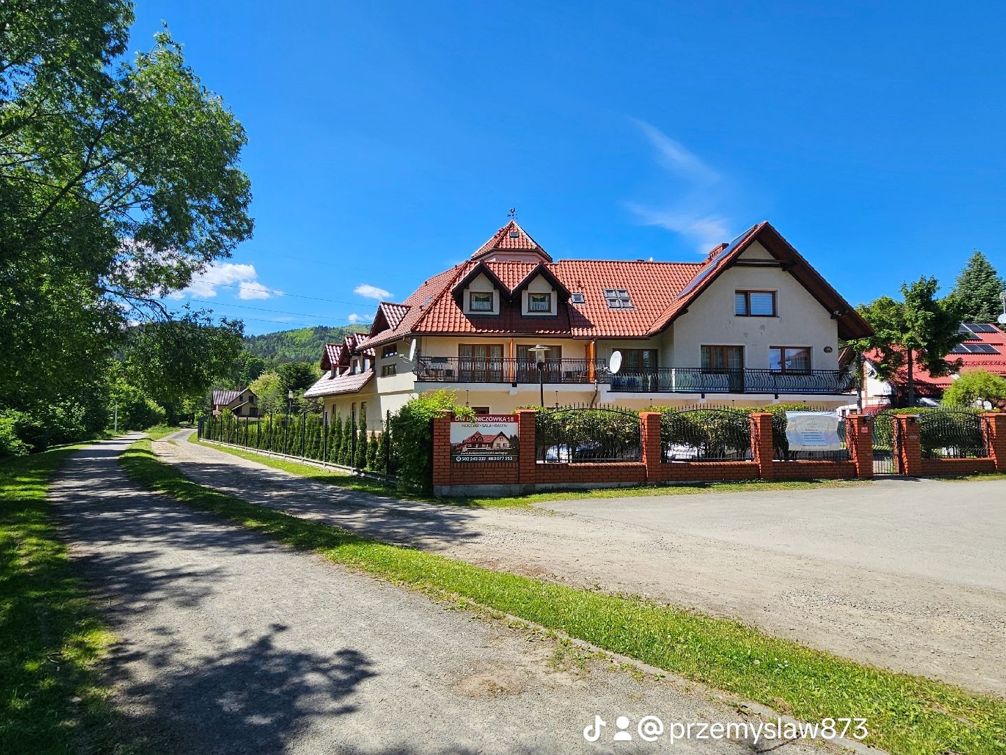 Dwupiętrowy pensjonat z czerwonym dachem i balkonami, otoczony zielenią i ogrodzeniem z cegły, widok z drogi gruntowej w słoneczny dzień.