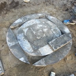 Mariusz Labuda - Układanie okręgu z szarych kostek brukowych na ziemi, widoczny gumowy młotek i buty robotnika, częściowo ułożony wzór geometryczny.