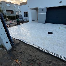 Mariusz Labuda - Nowoczesny podjazd z jasnoszarej kostki brukowej ułożonej przed minimalistycznym domem z czarną bramą garażową; widoczny pracownik w roboczym ubraniu i narzędzia w tle.