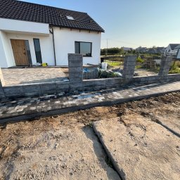 Mariusz Labuda - Wznoszona konstrukcja ogrodzenia z szarych, betonowych bloczków przy nowo wybudowanym, białym domu z szarą kostką brukową i przygotowanym podłożem.