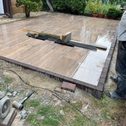 Mariusz Labuda - Układanie podłogi tarasu z płytek imitujących drewno, widoczne narzędzia i elementy pomocnicze, obrzeże z ciemnej kostki, etap prac, w tle zieleń ogrodu.