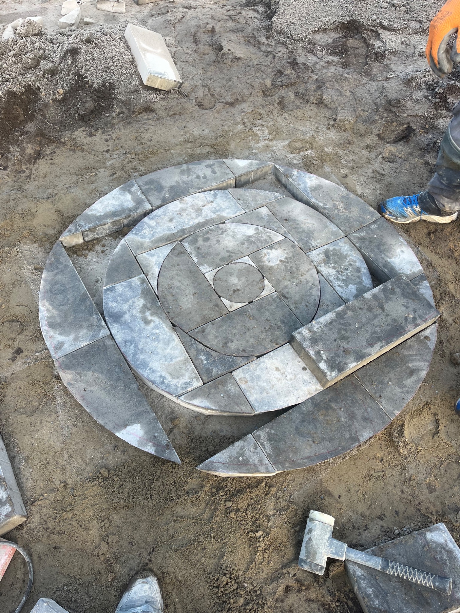 Układanie okręgu z szarych kostek brukowych na ziemi, widoczny gumowy młotek i buty robotnika, częściowo ułożony wzór geometryczny.
