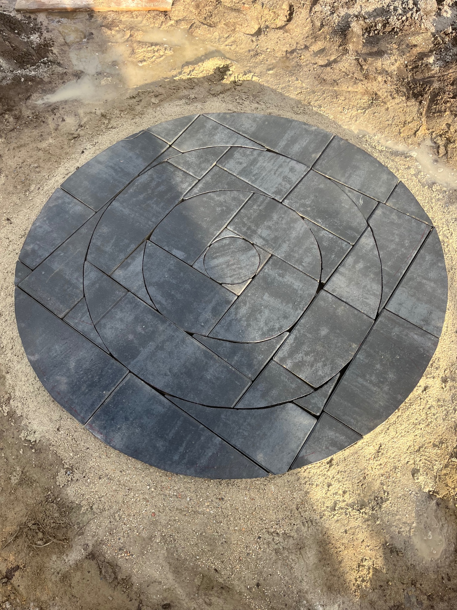 Okrągła kompozycja z szarych kostek brukowych ułożona na piasku, tworząca spiralny wzór w trakcie realizacji.