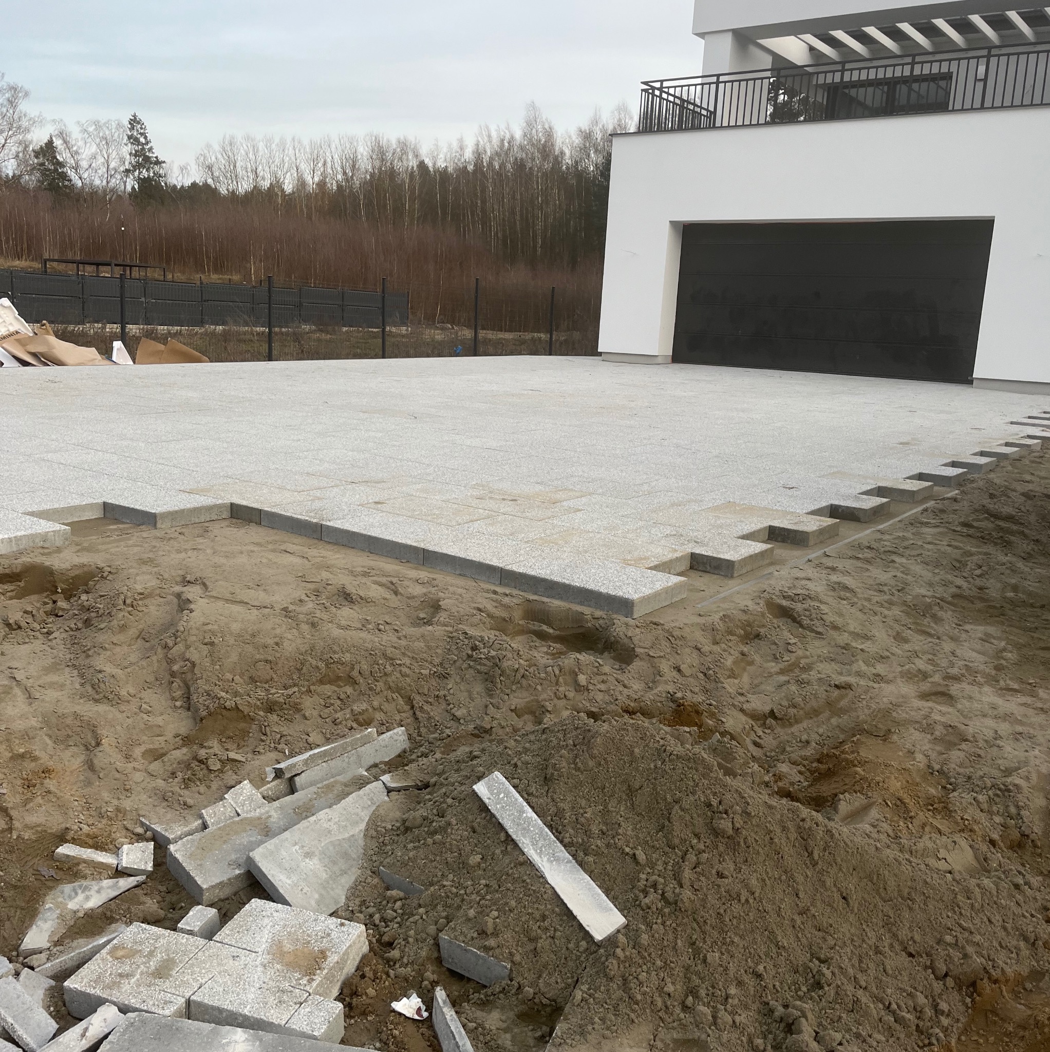 Układanie jasnoszarej kostki granitowej na podjeździe przed nowoczesnym domem z czarną bramą garażową, widoczne przygotowane podłoże i stosy kostki.