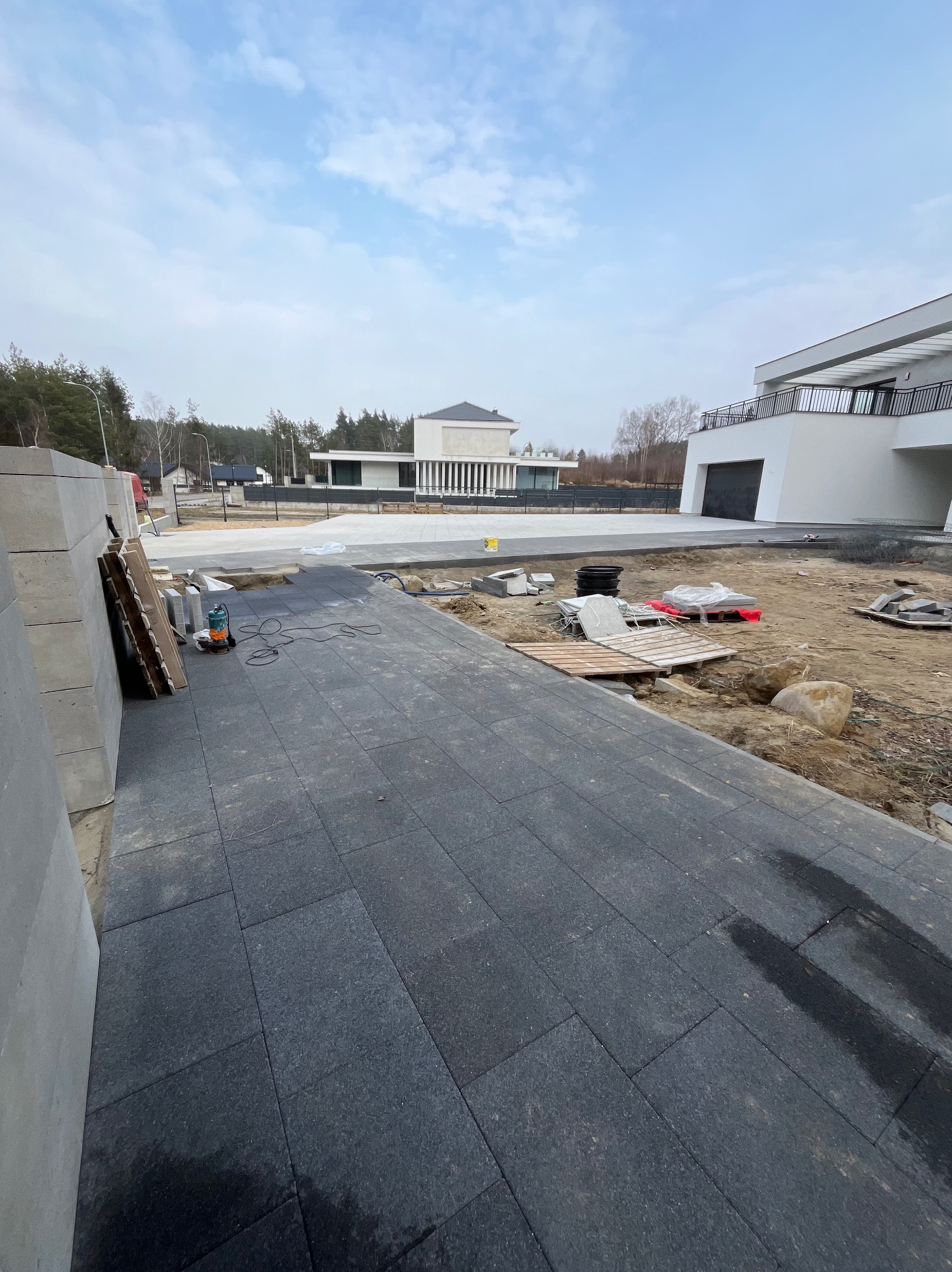 Częściowo ułożona, ciemnoszara kostka brukowa na podjeździe nowoczesnego domu. Widoczne narzędzia, palety i pozostałości materiałów budowlanych. W tle dwa budynki mieszkalne.