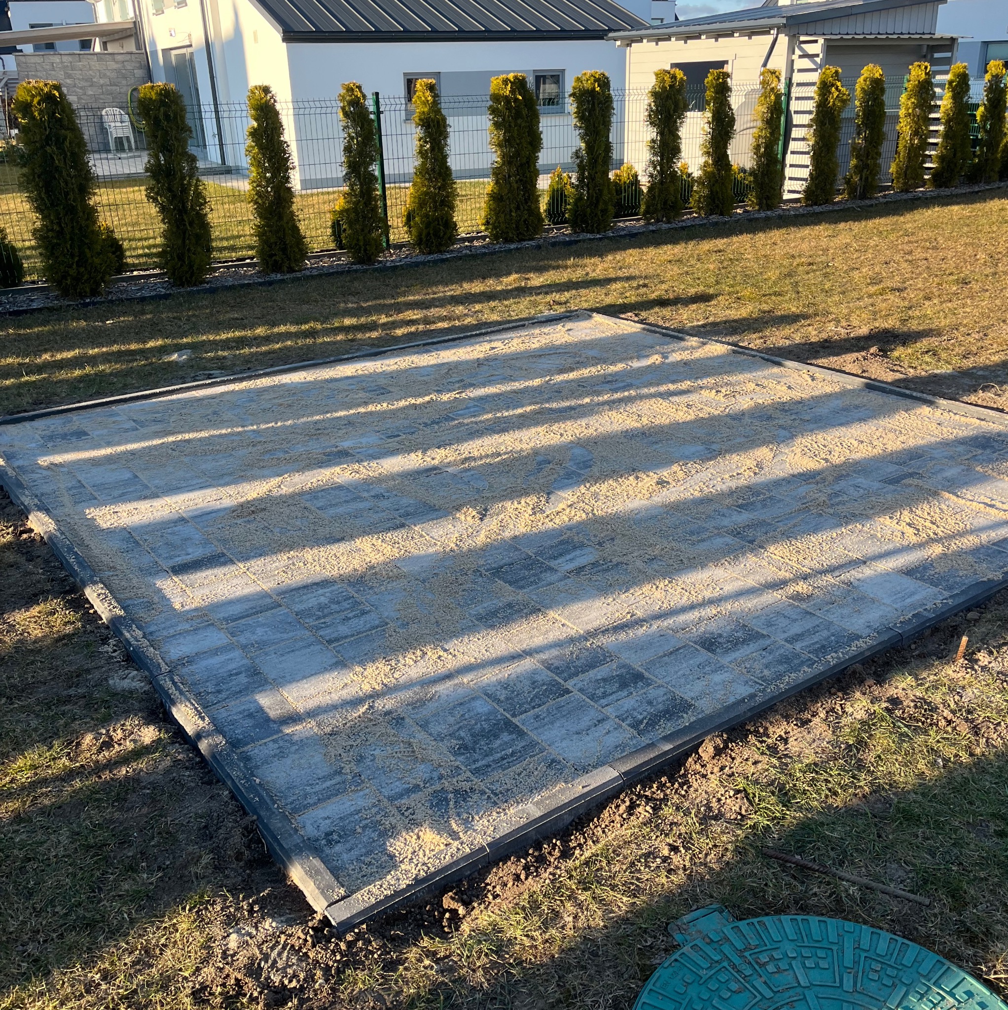 Przygotowany teren pod nawierzchnię z kostki brukowej, wysypany piaskiem i wyrównany, widoczne obrzeża, w tle rząd wysokich tuj.