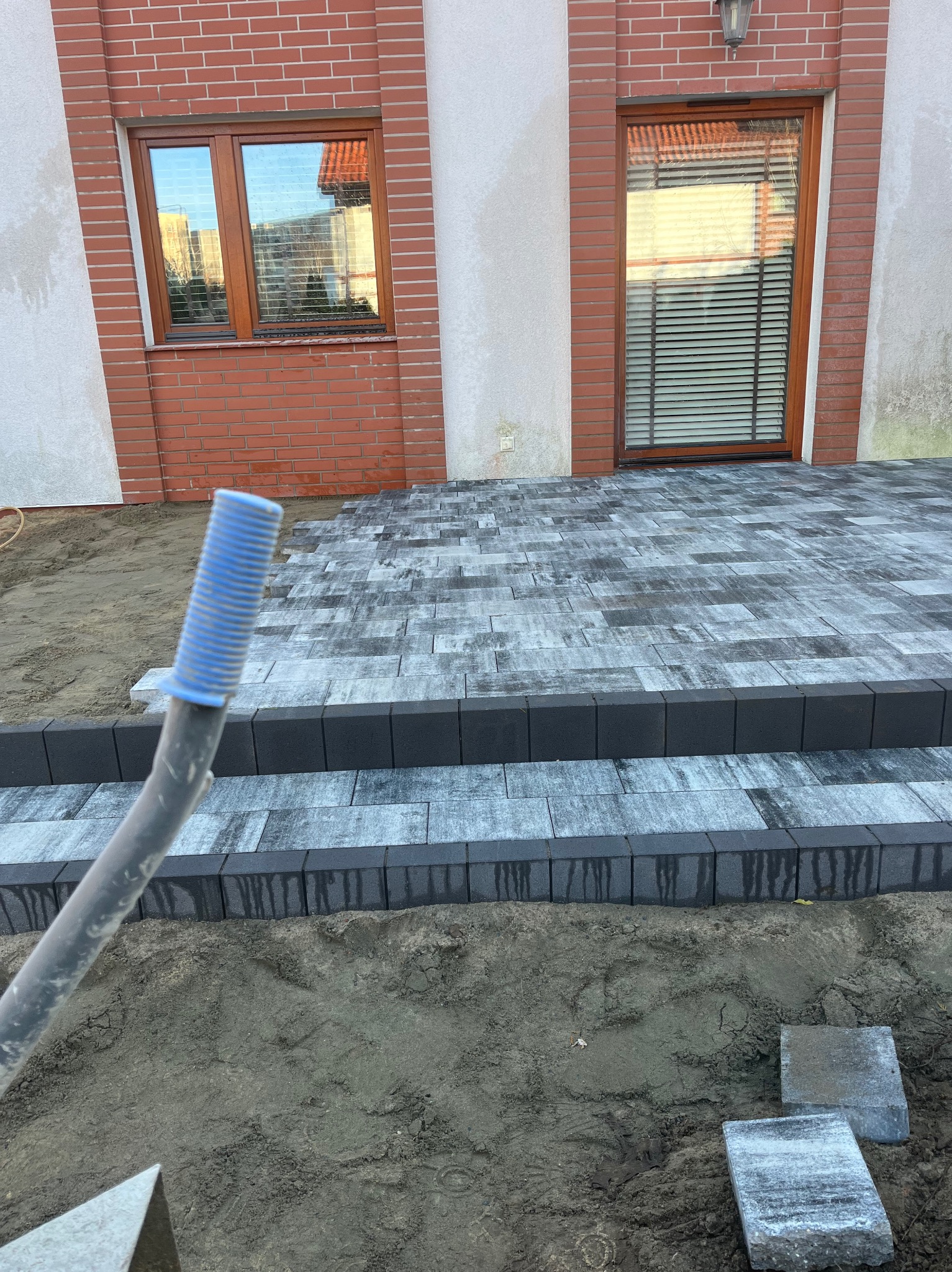 Ułożona kostka brukowa w odcieniach szarości, tworząca schody przed budynkiem z ceglaną elewacją i drewnianymi oknami. Częściowo widoczne narzędzie z niebieską końcówką. Praca w toku, widoczny piasek.
