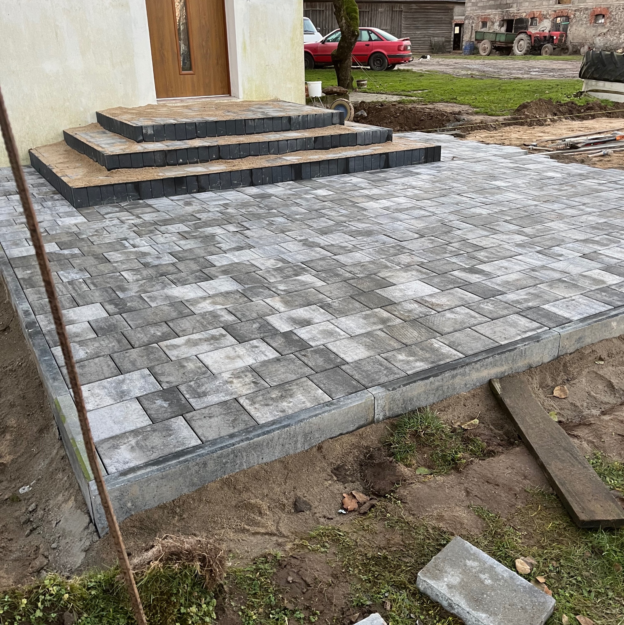 Nowo ułożona szara kostka brukowa prowadząca do stopni wejściowych domu, widoczne obrzeża i przygotowany teren pod dalsze prace brukarskie.