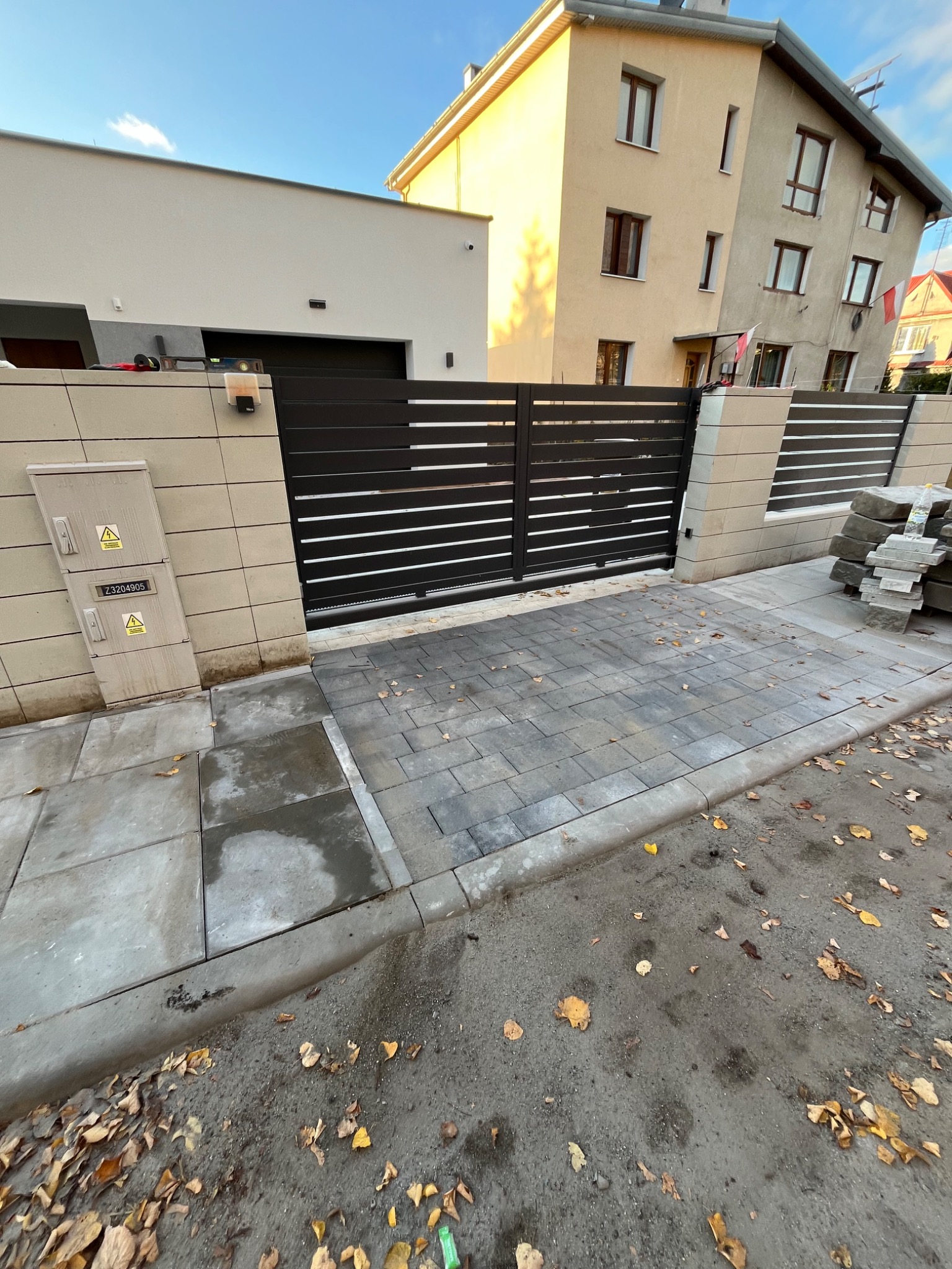 Nowoczesna posesja z przesuwną bramą z poziomych profili, wyłożona szarą kostką brukową, z widoczną skrzynką elektryczną na murze i stertą kostki obok