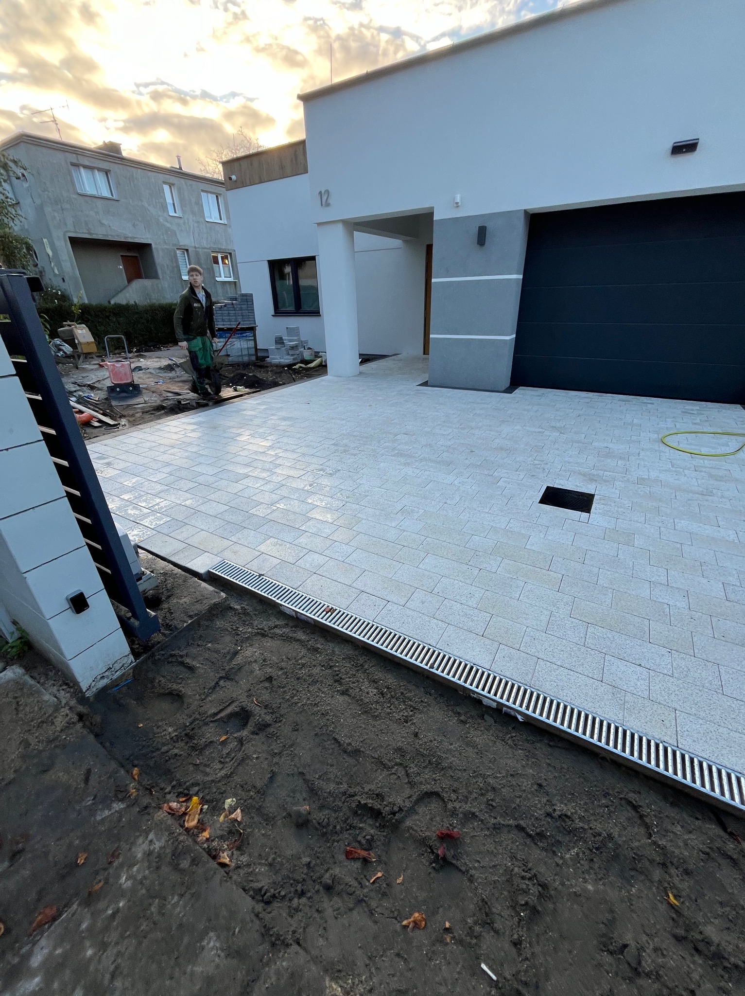 Nowoczesny podjazd z jasnoszarej kostki brukowej ułożonej przed minimalistycznym domem z czarną bramą garażową; widoczny pracownik w roboczym ubraniu i narzędzia w tle.