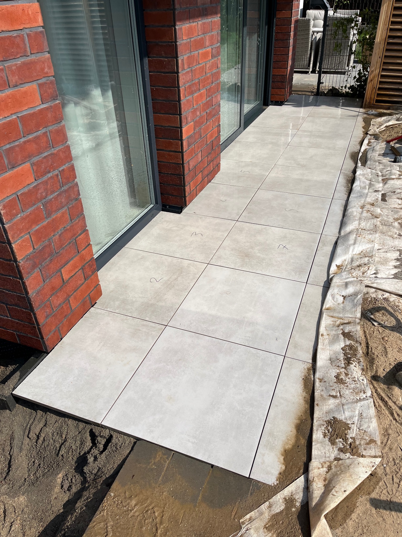 Jasnoszare płytki tarasowe ułożone przy ceglanej ścianie budynku. Widoczne ślady fugowania i zabrudzenia budowlane.