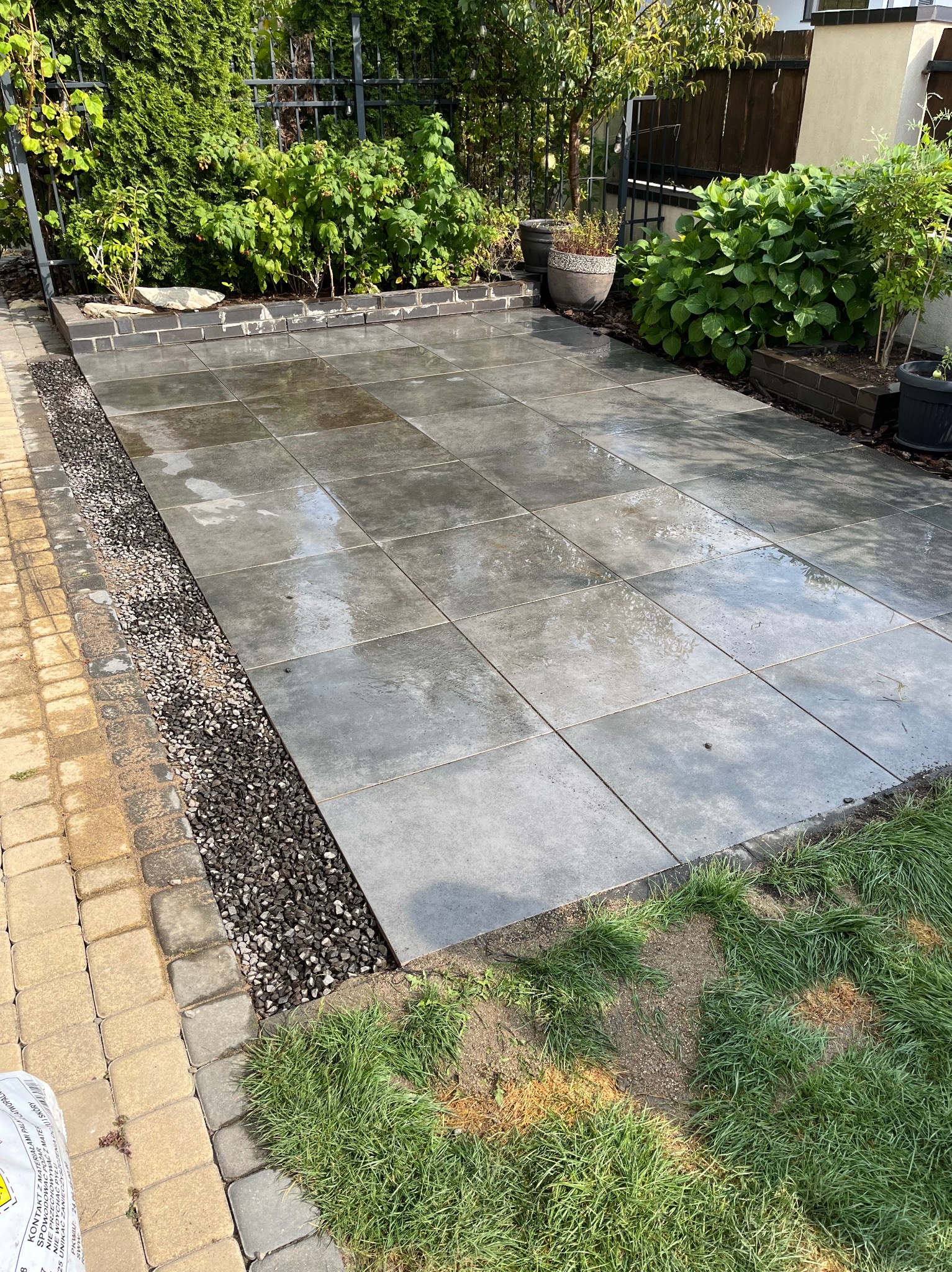 Świeżo ułożony, mokry taras z szarych płyt betonowych, oddzielony od trawnika pasem czarnego grysu, obrzeże z kostki brukowej w dwóch kolorach, zieleń w tle.