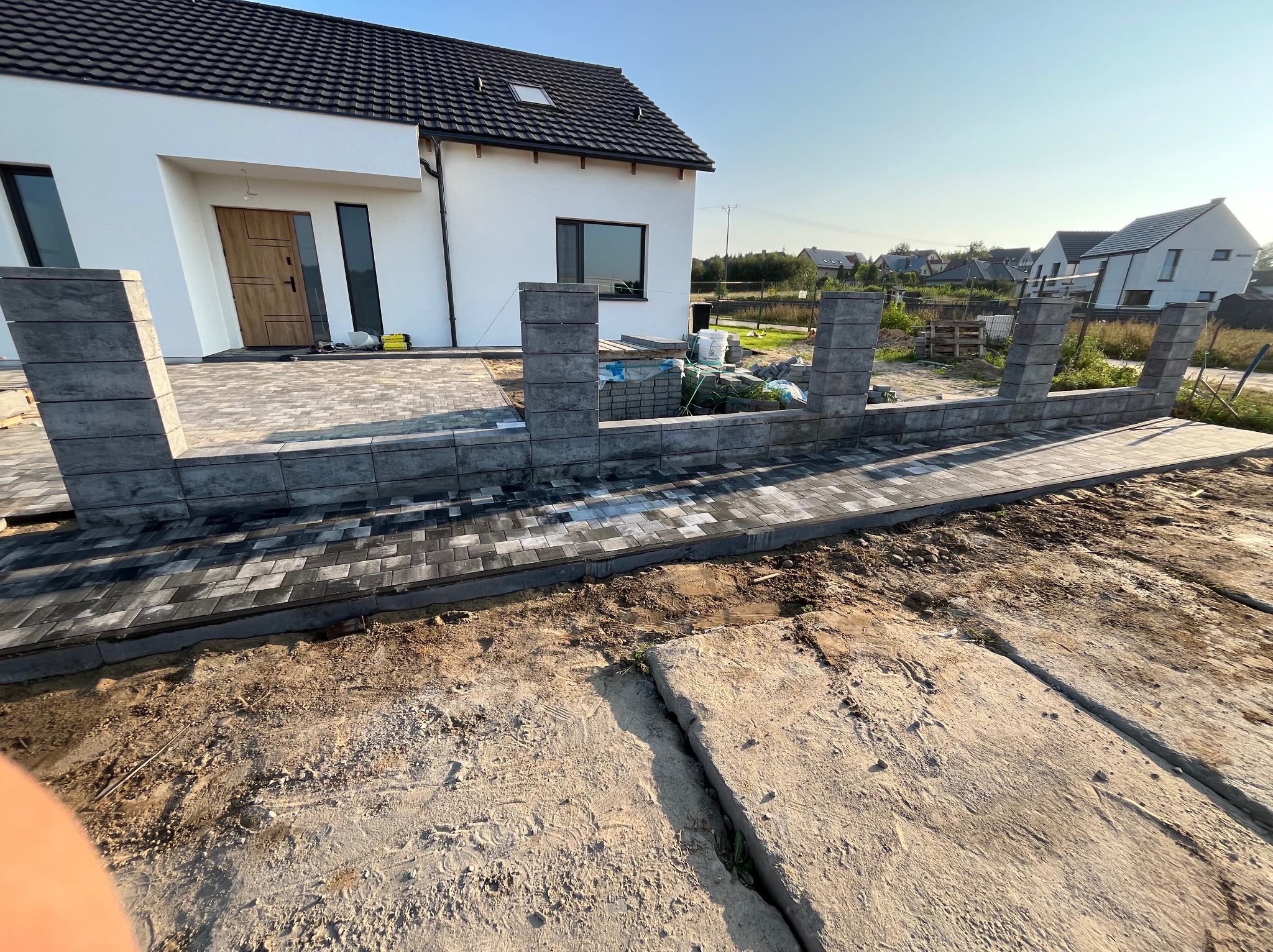 Wznoszona konstrukcja ogrodzenia z szarych, betonowych bloczków przy nowo wybudowanym, białym domu z szarą kostką brukową i przygotowanym podłożem.